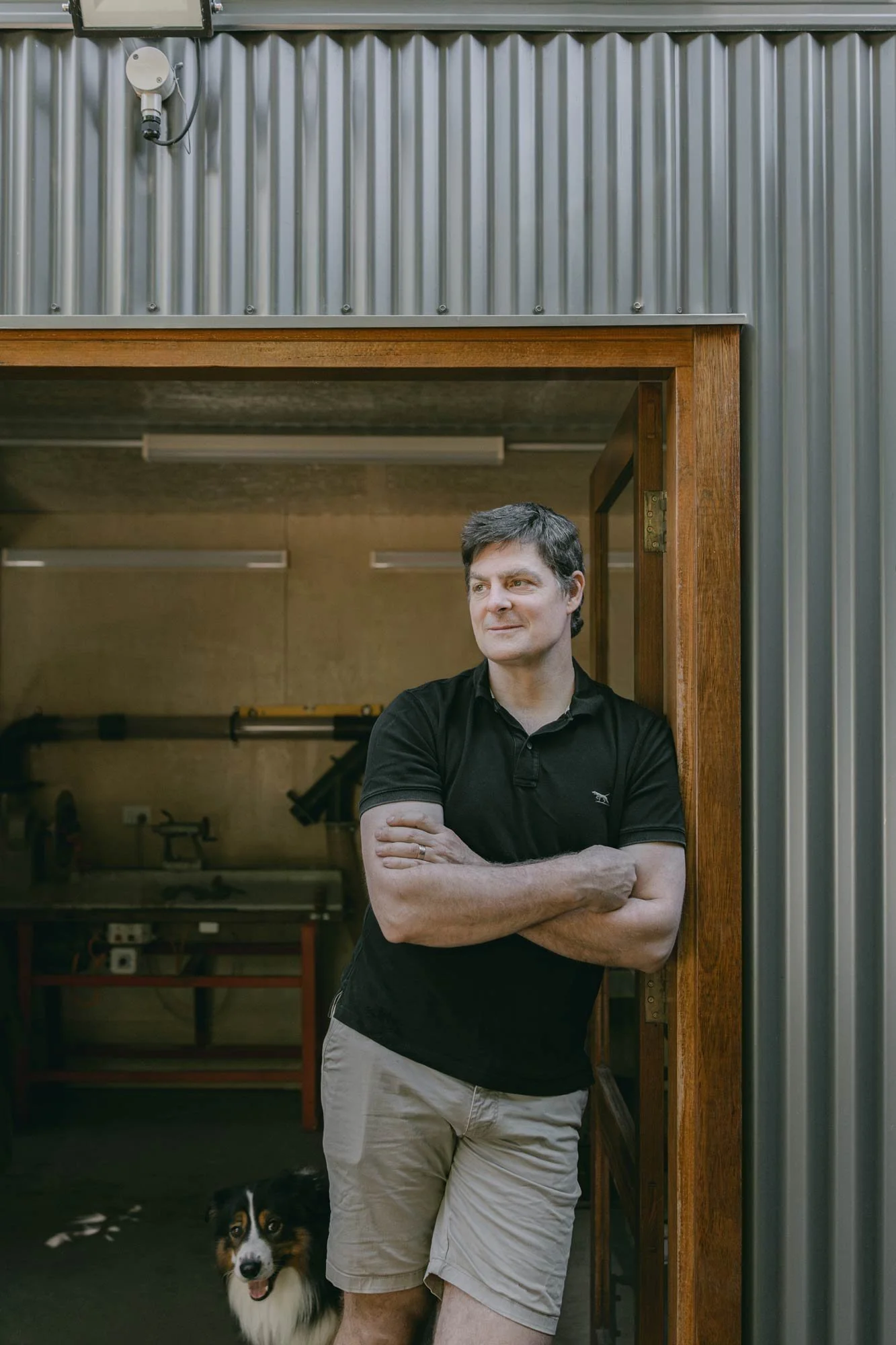 Australian wood turner, Andrew McNamara, with crossed arms standing in front of the doorway to his woodworking workshop, with his dog at his feet.