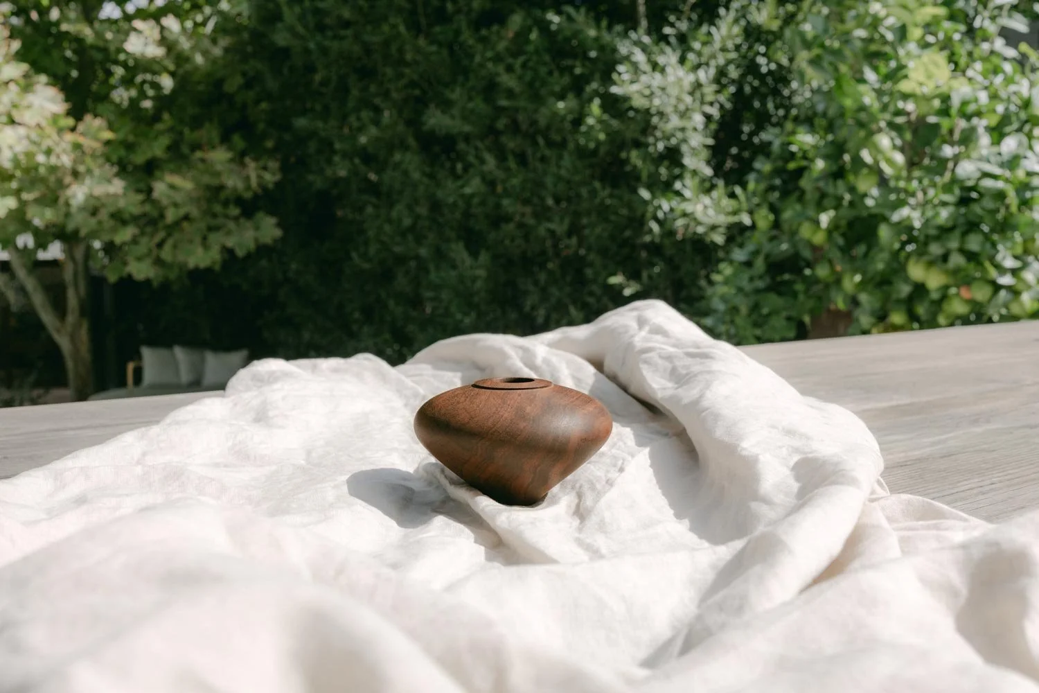 A small wooden vase placed on white fabric with a background of green trees and bushes.