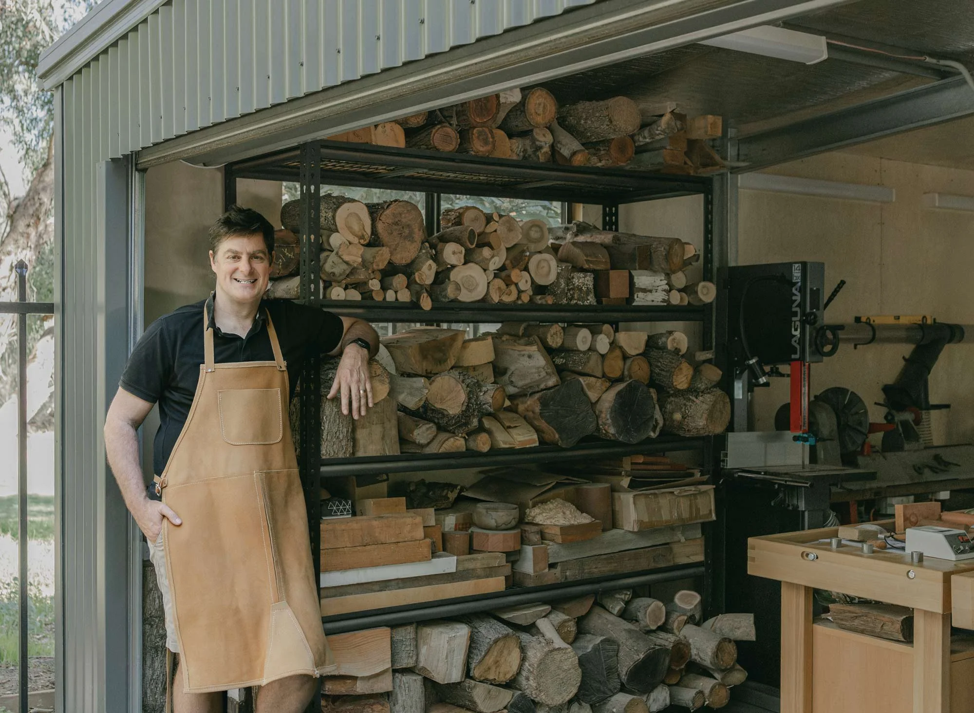 Australian wood turner, Andrew McNamara, wearing a tan apron and black shirt standing beside a metal firewood rack filled with cut logs in his woodturning workshop.