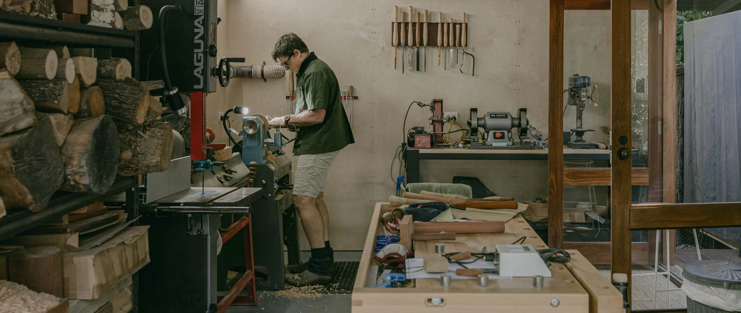Andrew McNamara working in his woodworking workshop, using a lathe machine. The workshop contains stacks of firewood on shelves, various tools hanging on the wall, and woodworking equipment on benches.