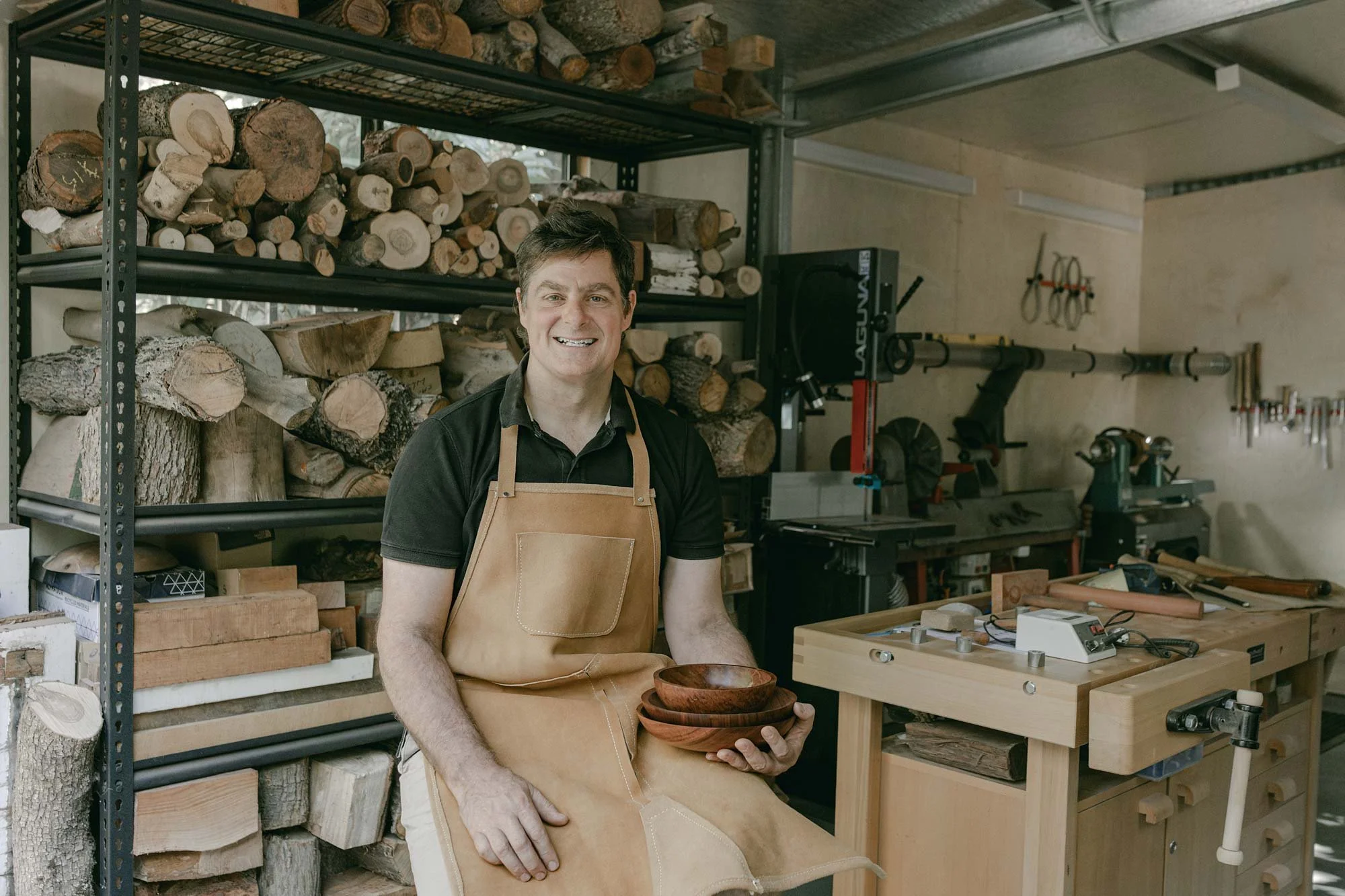 Australian wood turner, Andrew McNamara, in his woodworking workshop holding wooden bowls, surrounded by shelves of logs and woodworking tools.