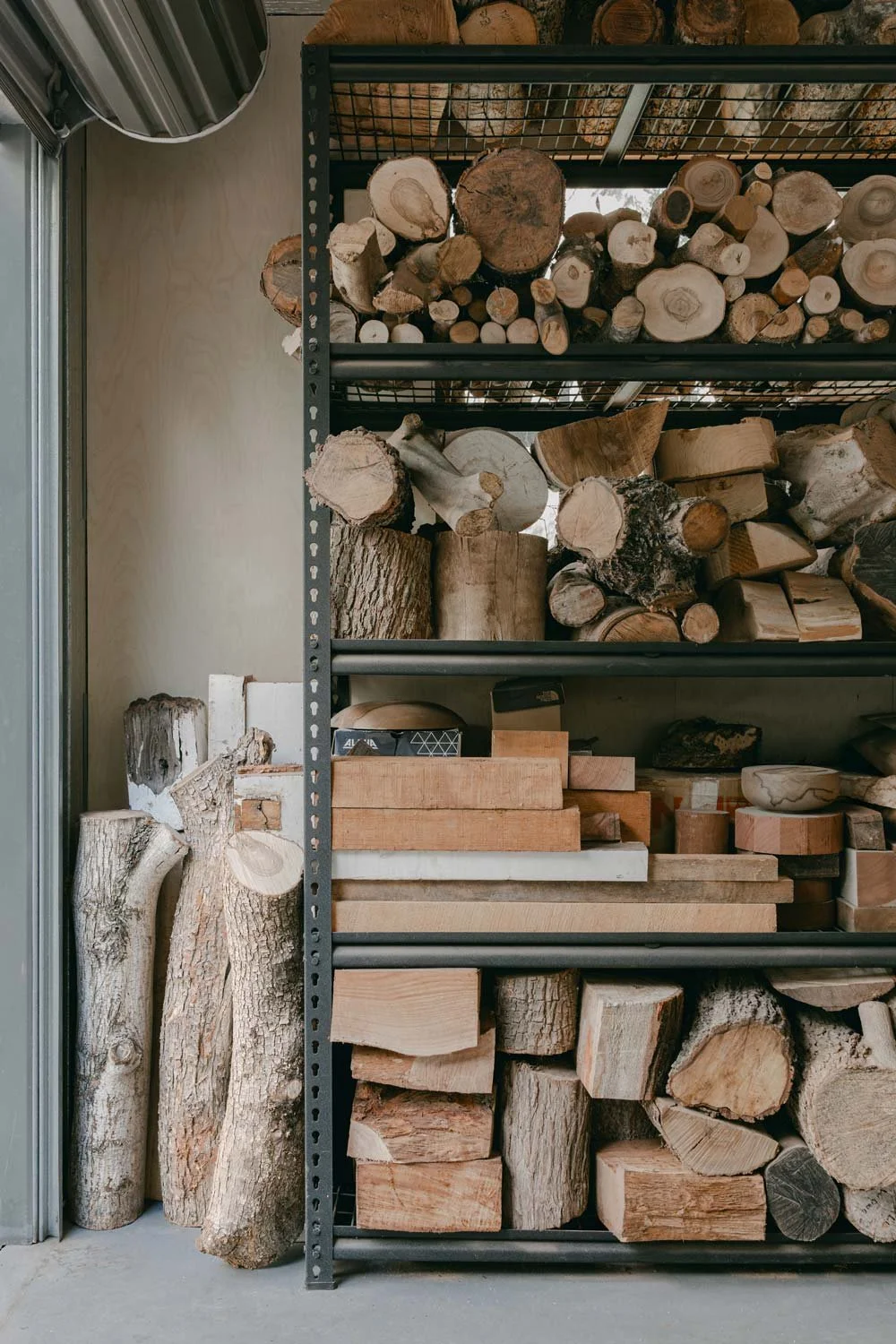 Wood logs stored on a metal shelving unit and some logs on the floor next to the shelving in Andrew McNamara Woodturning workshop.