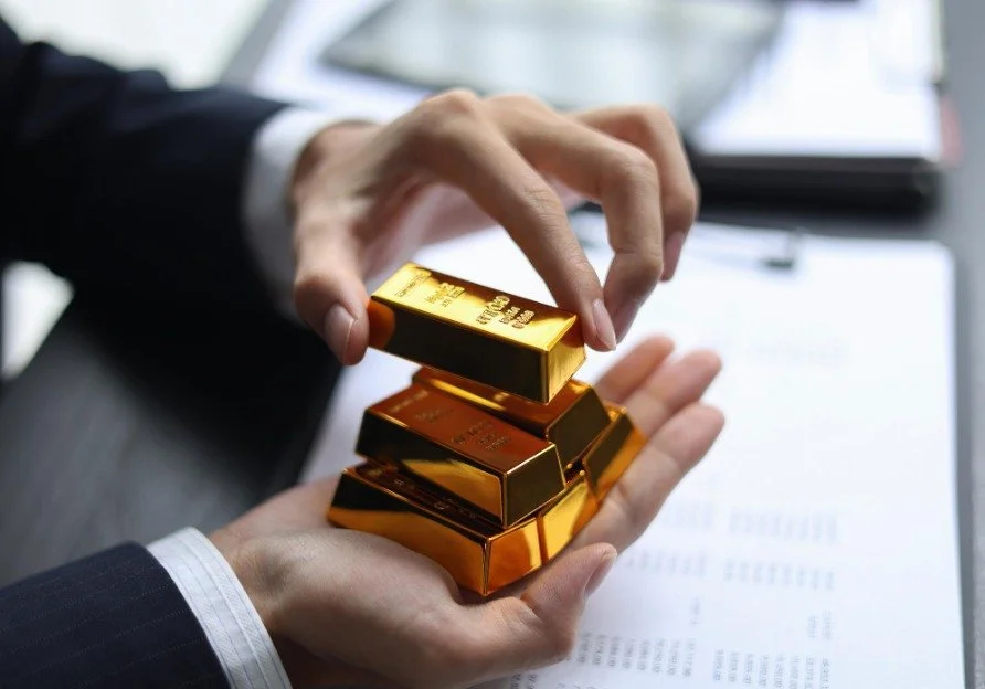 Person in business attire stacking gold bars on a dark table with a document and pen nearby.