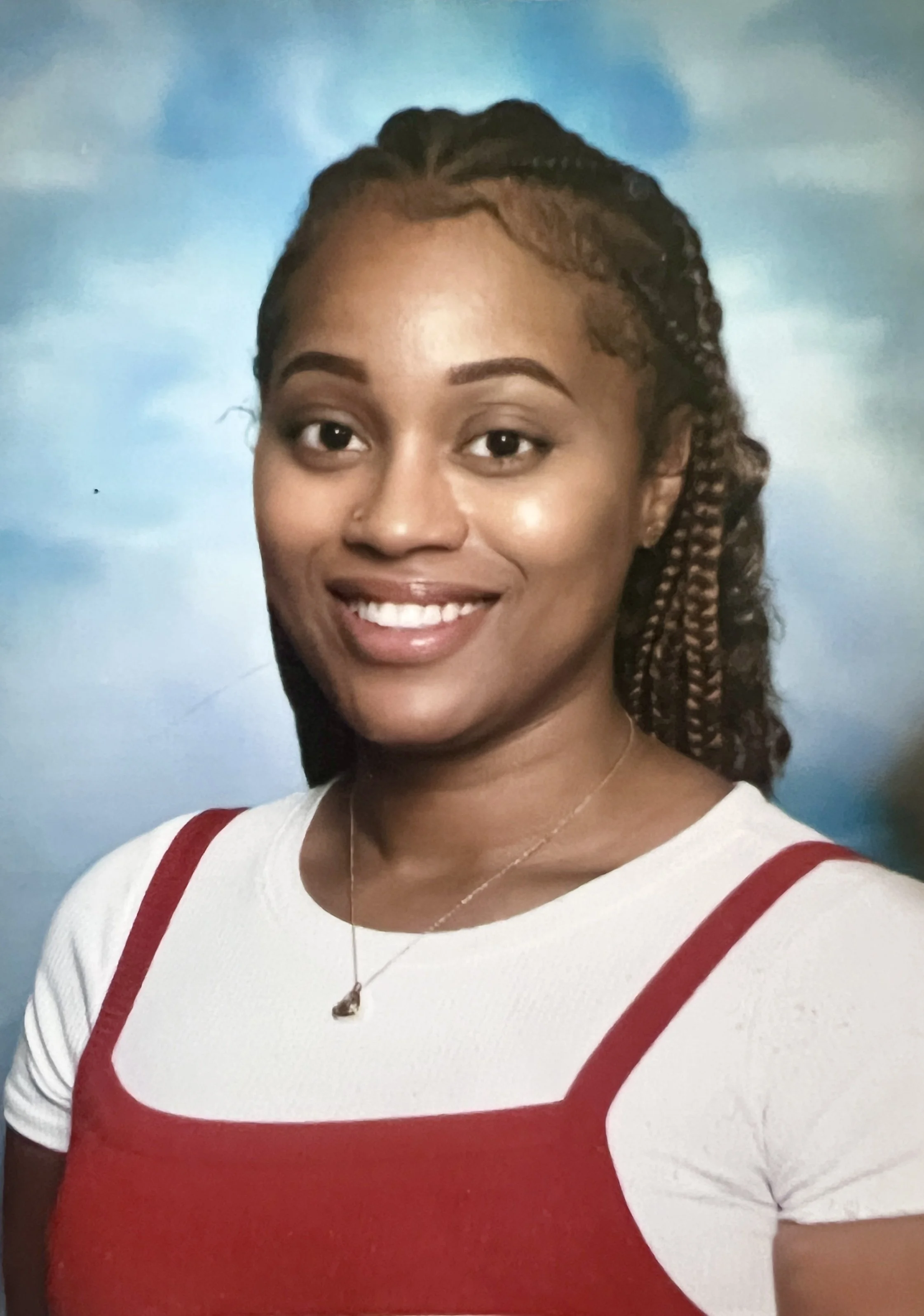 A smiling woman with braided hair wearing a white shirt with red straps, a pearl necklace, and a small pendant, standing against a blue sky background.