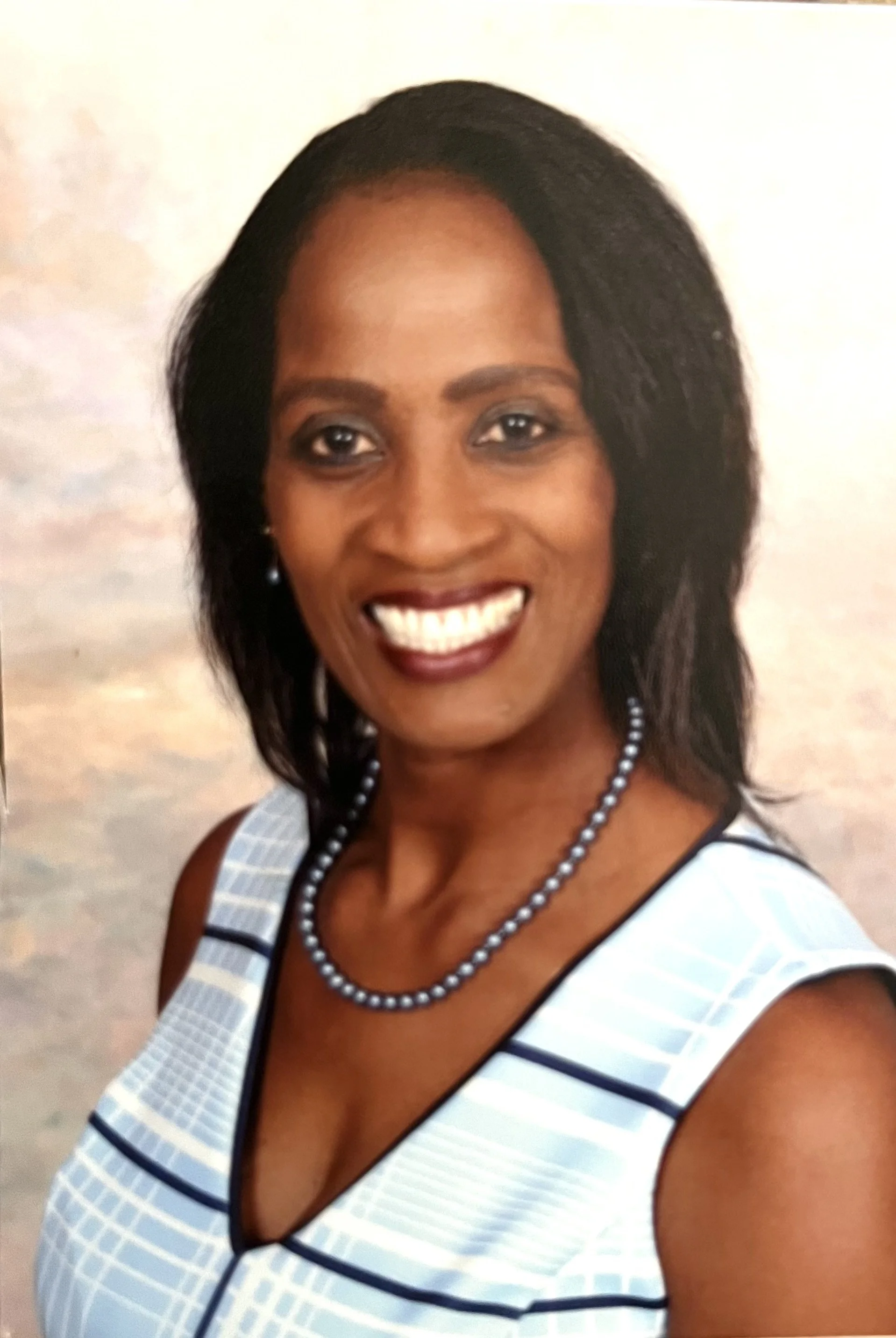 Close-up photo of a smiling woman with medium-dark skin, black hair, wearing a white and blue sleeveless dress with black piping, a pearl necklace, and pearl earrings.