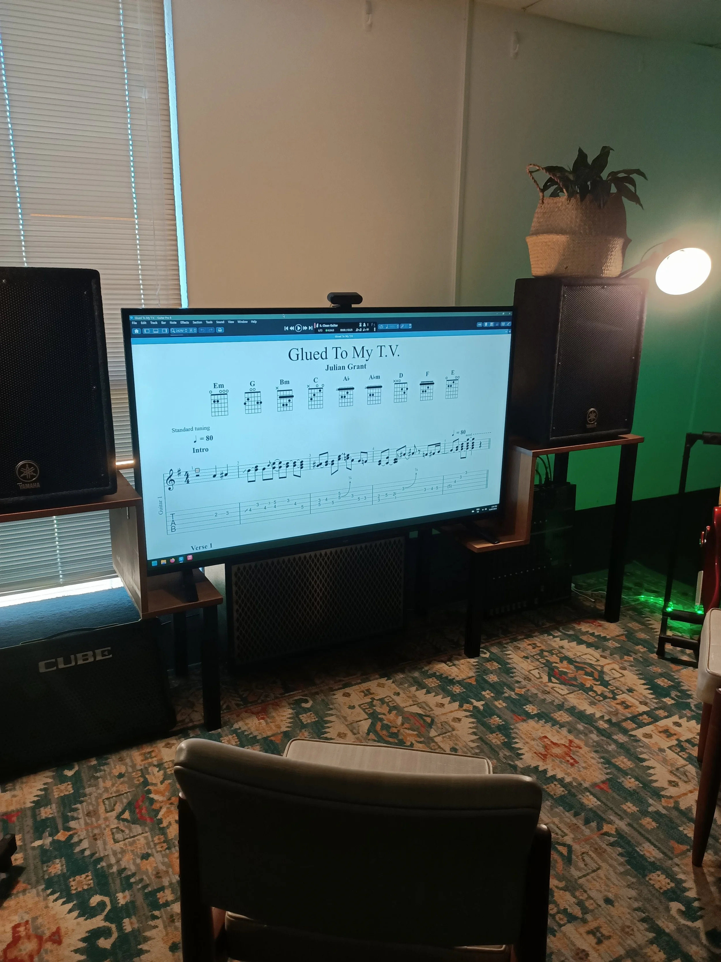 A computer monitor on a desk displaying guitar chords and sheet music for the song 'Glued To My T.V.' by Julian Grant. Two black speakers are on either side of the monitor. The room has a patterned carpet, a window with closed blinds, a potted plant 