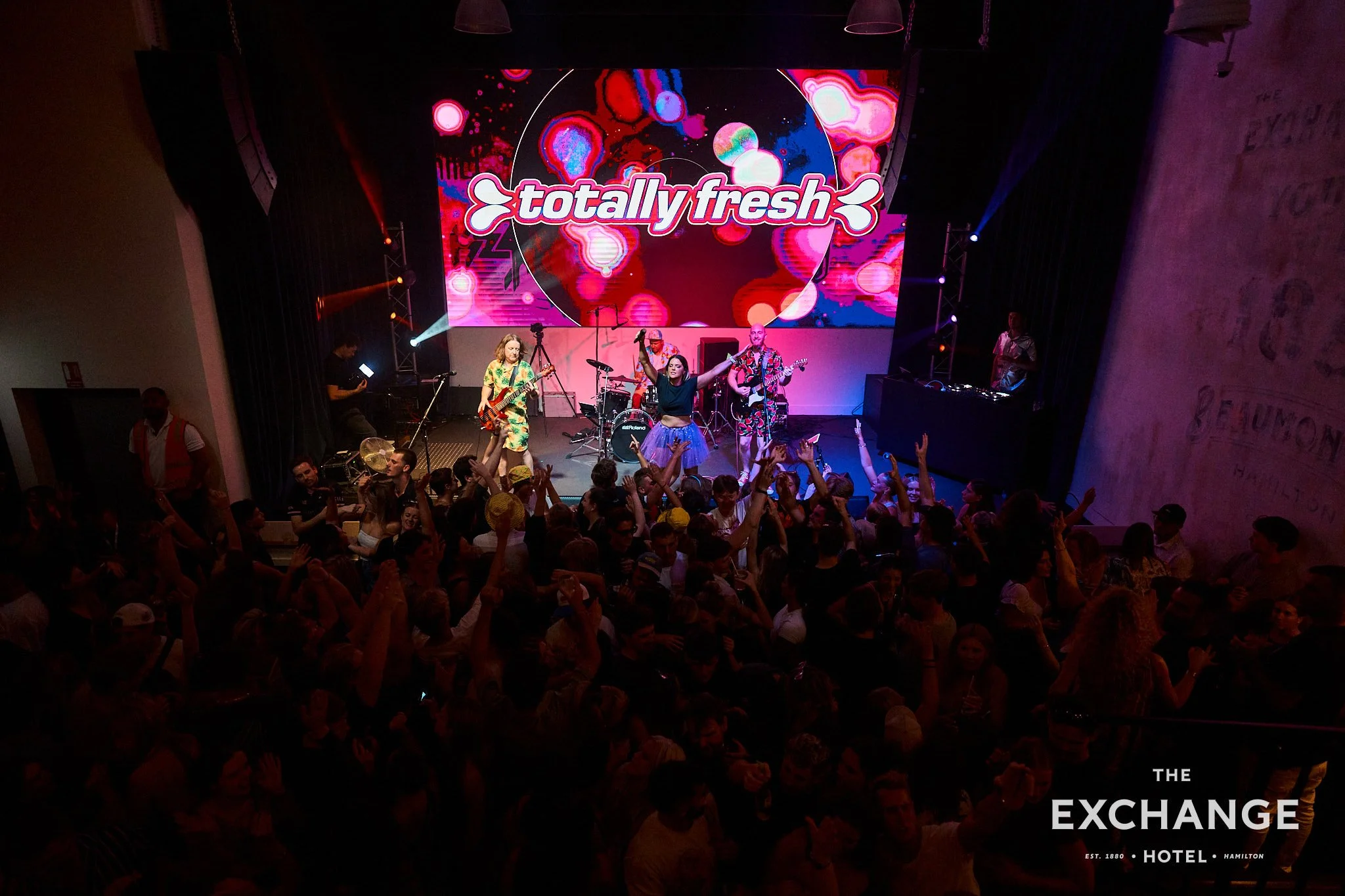 A live music performance at a crowded event with a large digital screen displaying the words 'totally fresh' and colorful abstract graphics. The band members are playing instruments on stage, and the audience is engaged.