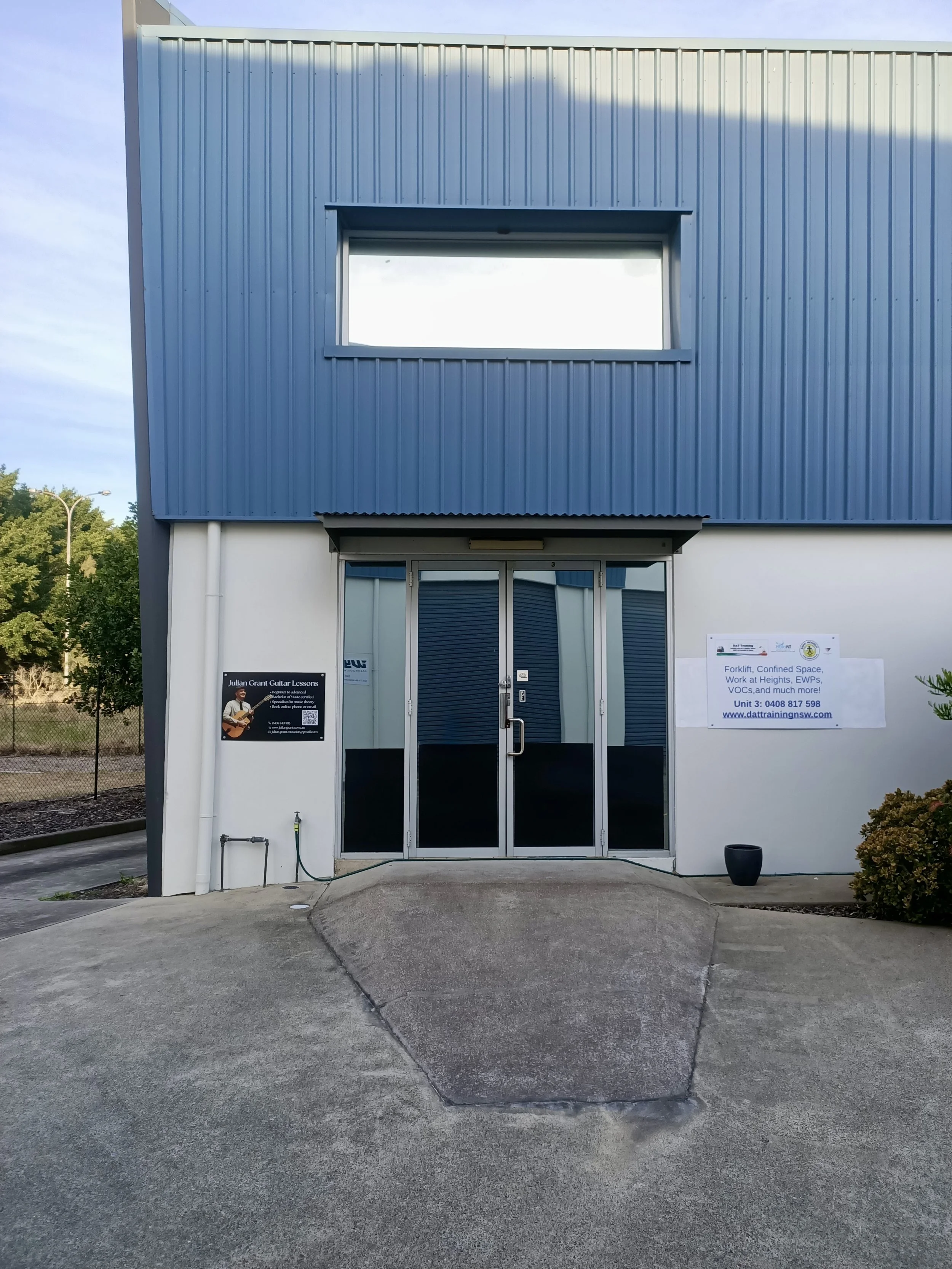 Image of a modern commercial building with a blue corrugated metal upper section and a white lower section. The entrance features glass doors with a sign to the left advertising guitar lessons and a larger sign to the right mentioning various trainin
