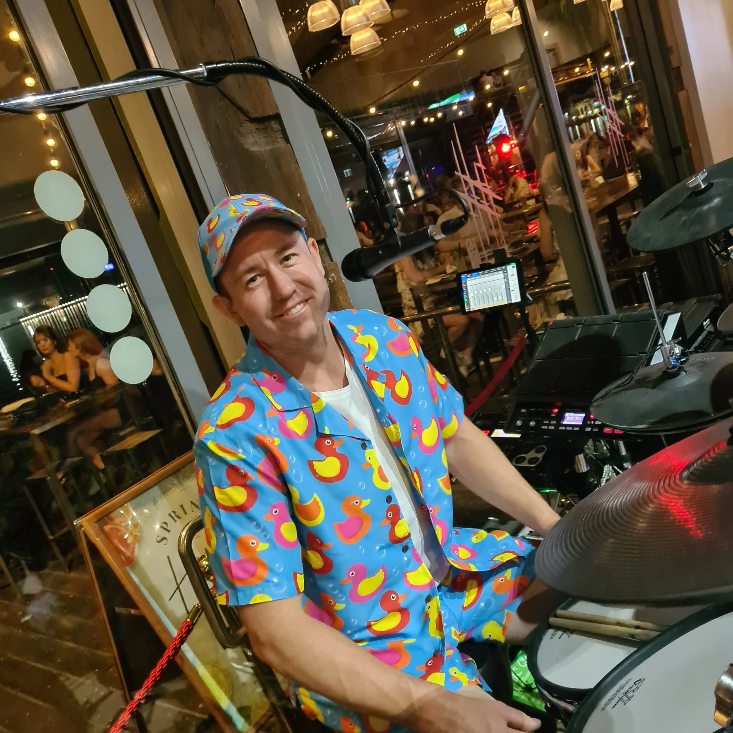 A man wearing a colorful hat and shirt with rubber duck patterns is sitting behind a drumkit at a bar or restaurant with string lights and large windows revealing a night cityscape. There are people in the background inside the venue.