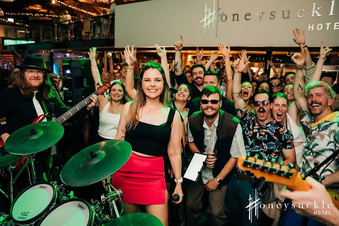 A lively indoor concert scene at Honeysuckle Hotel with a band performing. A smiling woman in a black top and red skirt holds a microphone in front of a drum set, surrounded by an energetic crowd with their hands raised, many wearing sunglasses and s