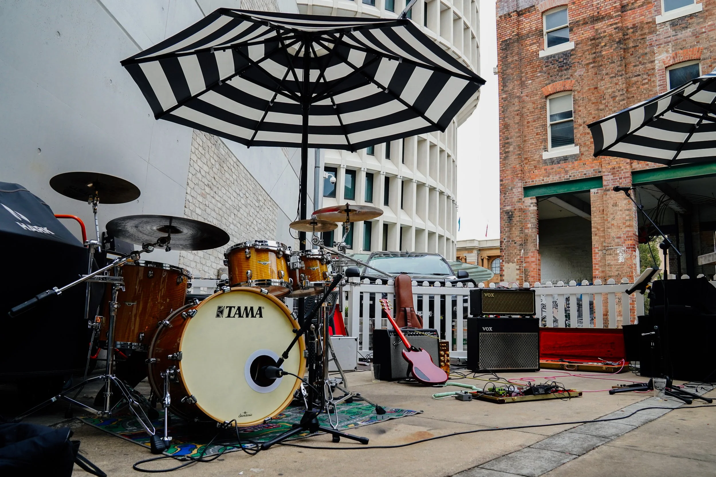 Musical instruments including a Tama drum set, guitars, and amplifiers set up outdoors on a stage with umbrellas and a backdrop of buildings.