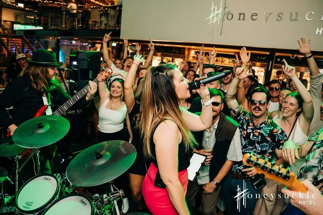Female singer performing with a microphone on stage at a lively indoor concert, with a crowd of enthusiastic people raising their hands and enjoying the music.