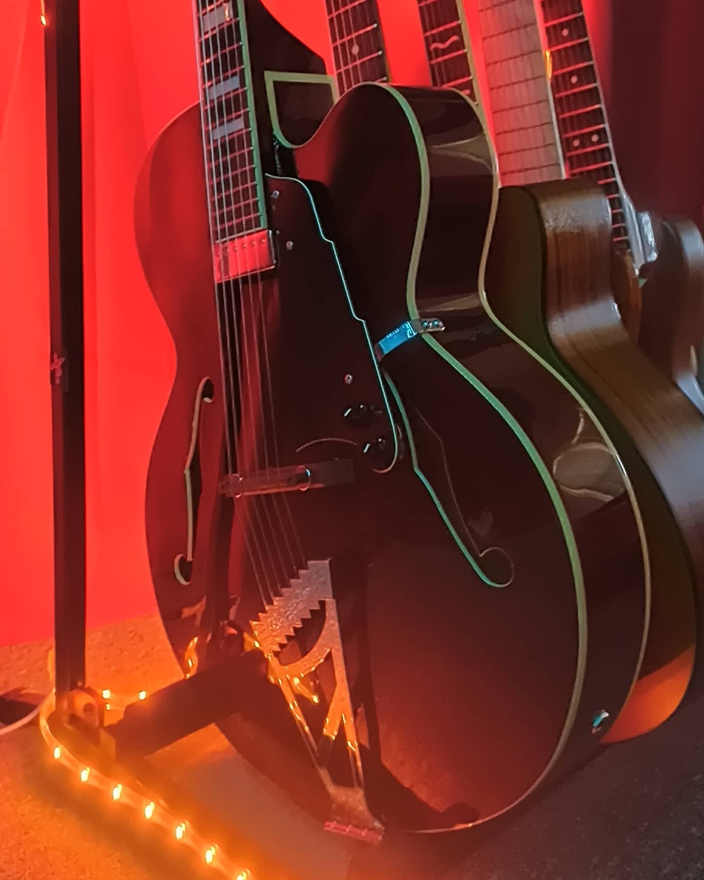 A black guitar with colorful neon-like trim, illuminated by orange string lights, on a stand.