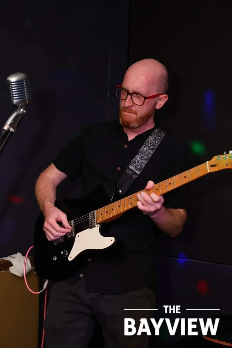 A man with glasses and a beard playing an electric guitar in a dark room, with a microphone nearby, and a logo reading 'The Bayview' at the bottom right corner.