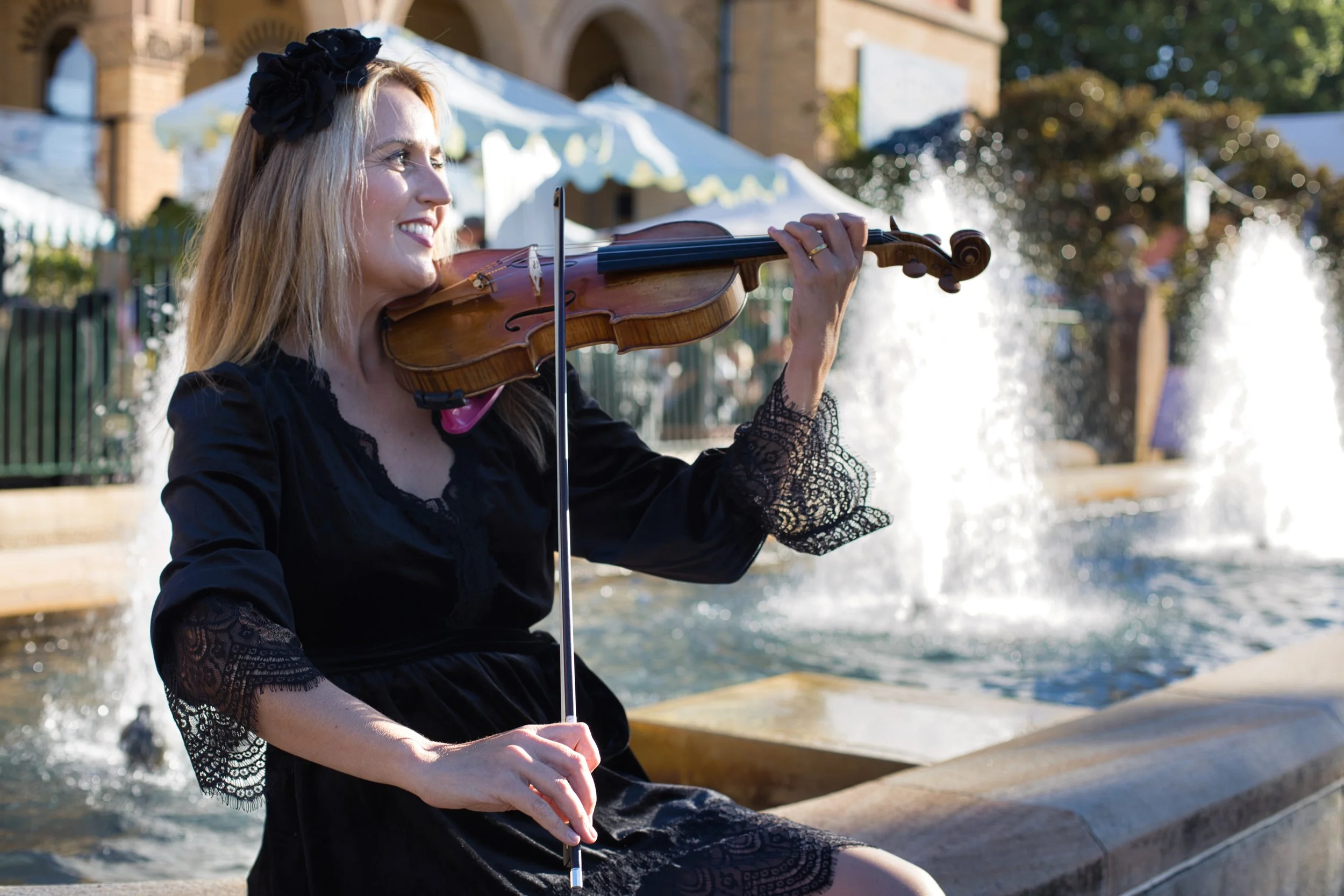 A woman with blonde hair wearing a black dress and a large black floral headband, playing the violin outdoors near a water fountain with a building and trees in the background.