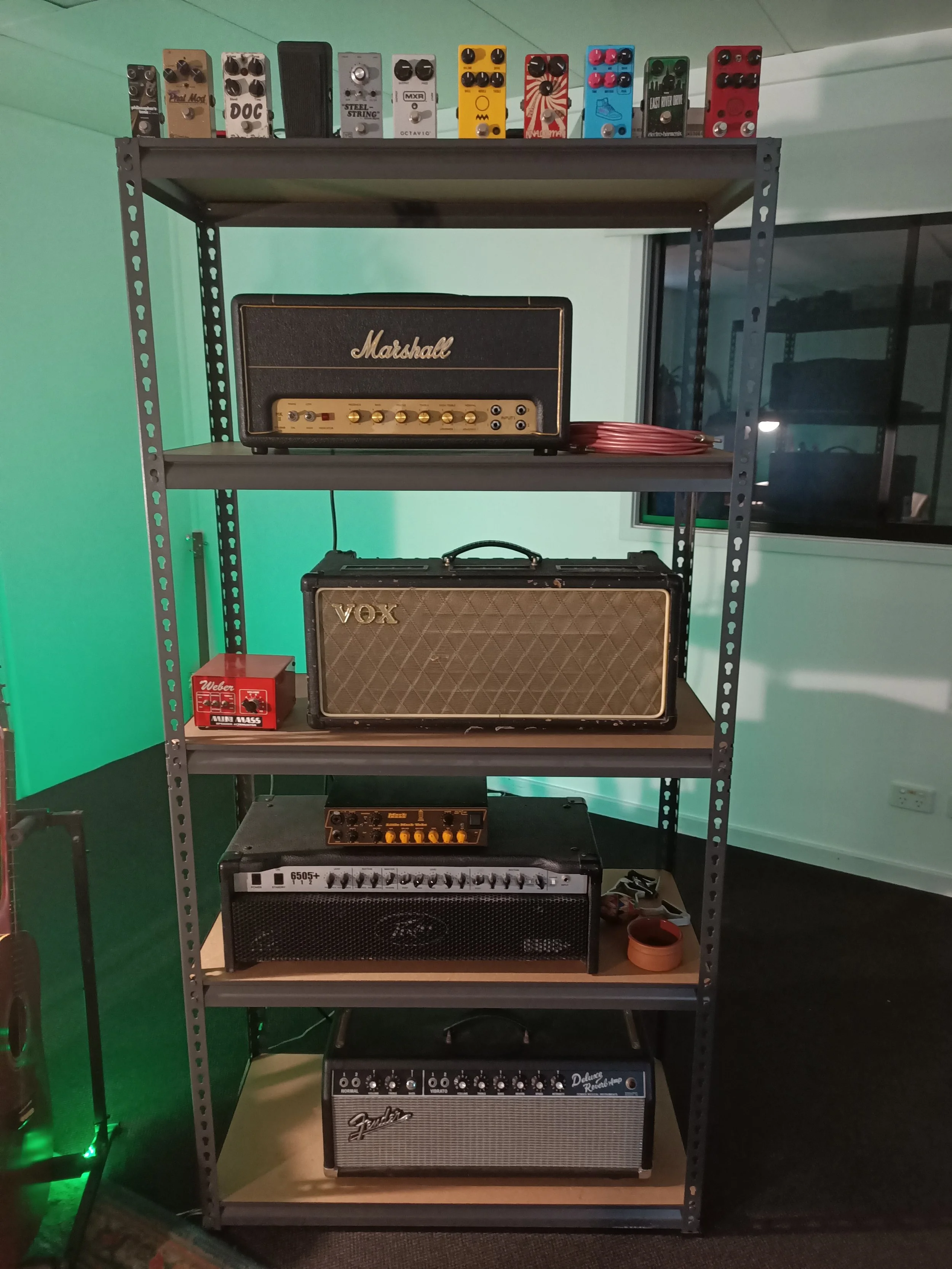 A metal shelving unit with guitar amplifiers and effects pedals. The top shelf has various guitar effect pedals, the second shelf has a Marshall guitar amplifier, the third shelf has a Vox guitar amplifier, and the bottom shelf has a Fender guitar am