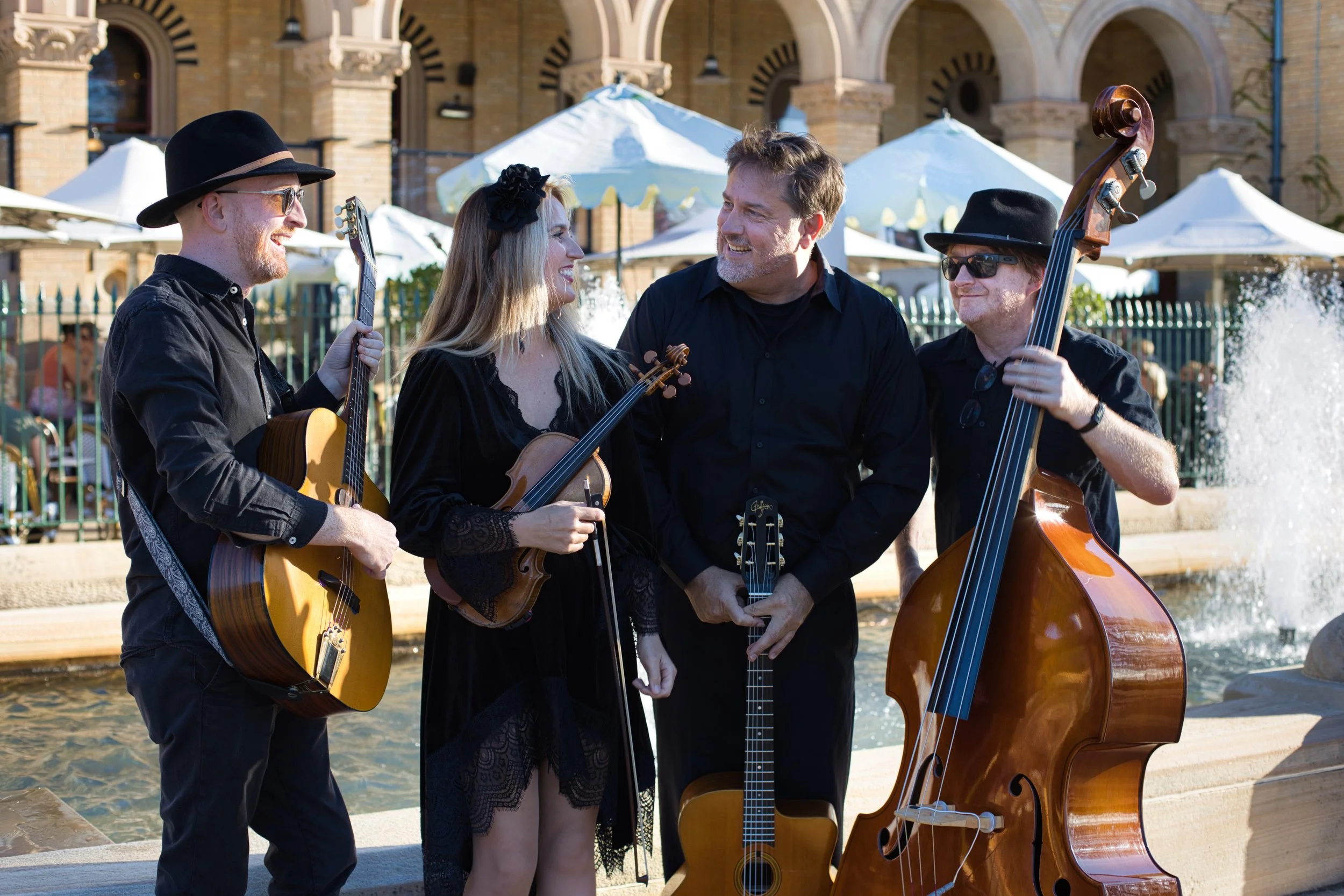Four musicians playing instruments near a fountain in an outdoor setting, with a building and white umbrellas in the background.