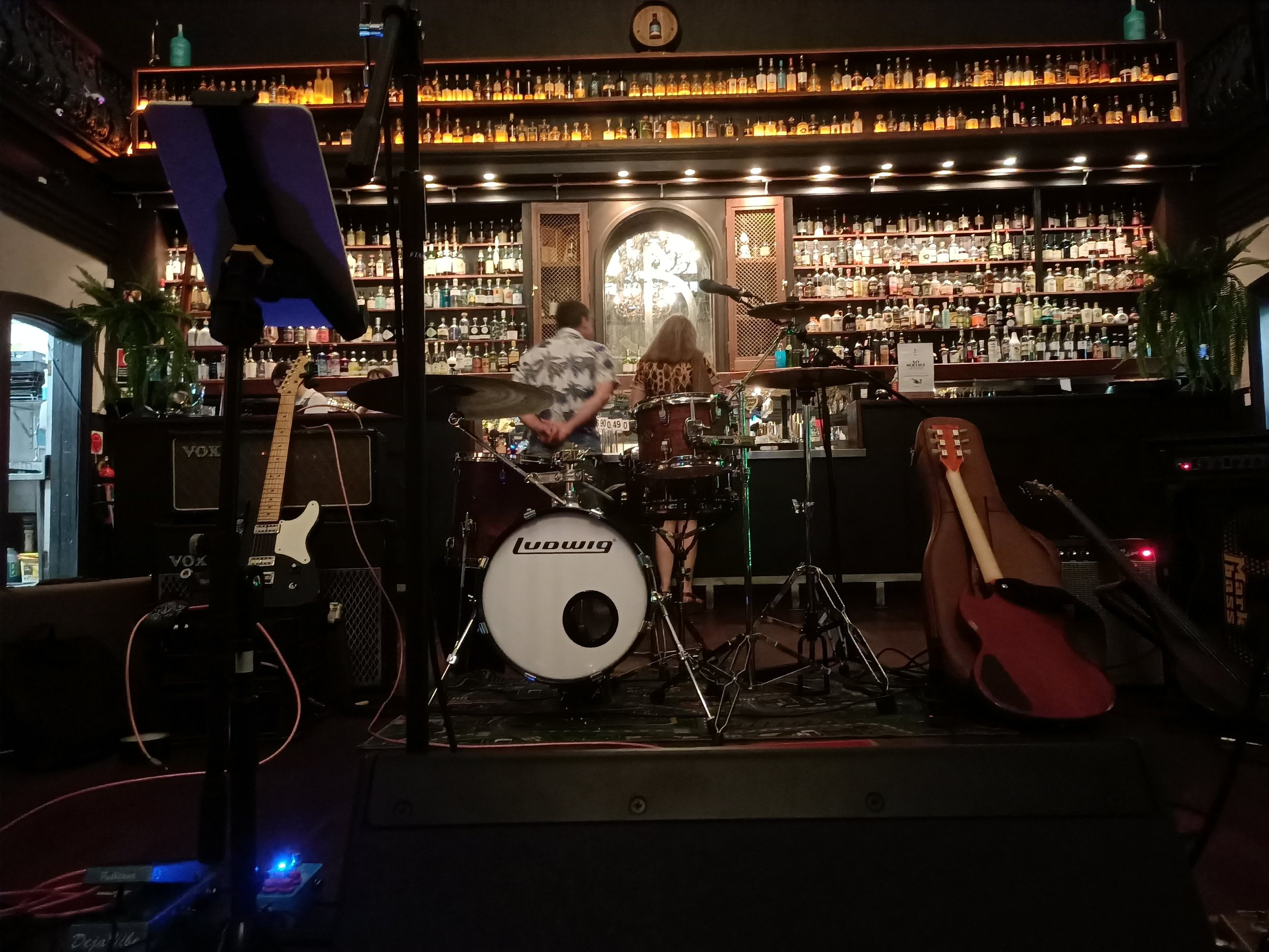 A dimly lit bar or music venue with shelves of liquor bottles on the wall behind a small stage. There is a drum set, an electric guitar, and music equipment on the stage. Two people are standing near the drum set, possibly preparing for a performance
