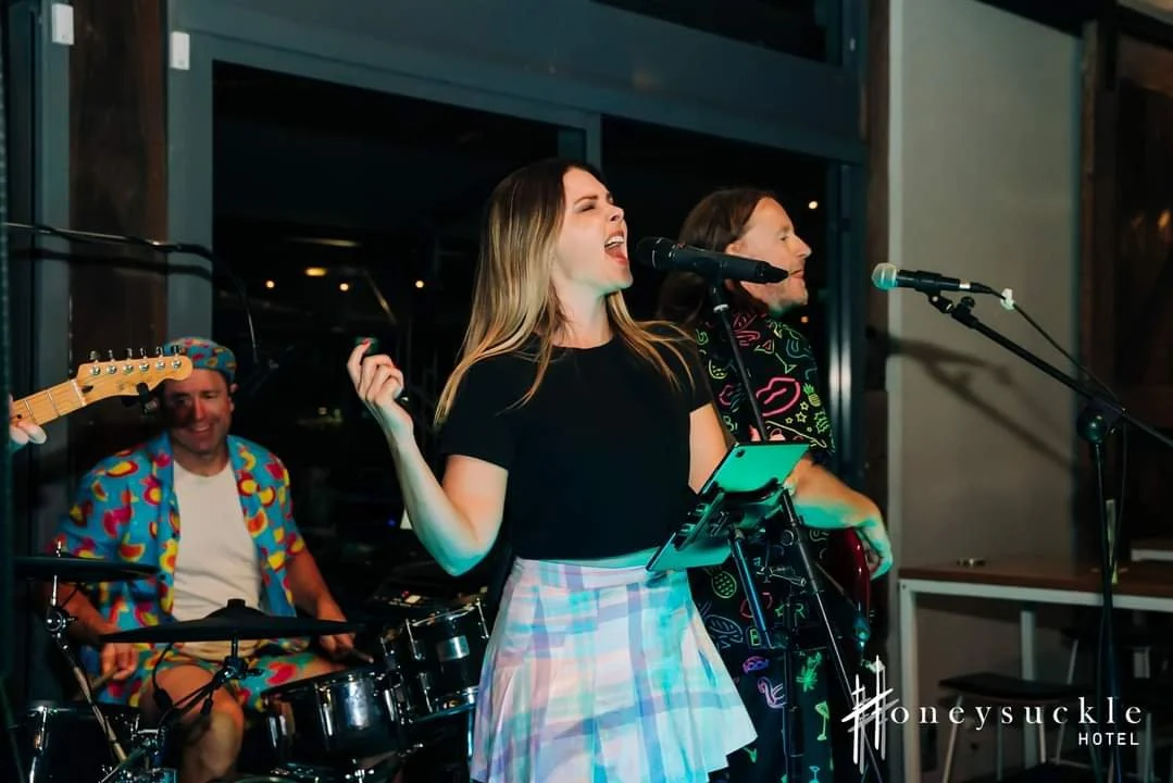 A woman singing into a microphone with a band performing behind her at an indoor live music event, with a drummer in a colorful shirt and a guitarist next to her.