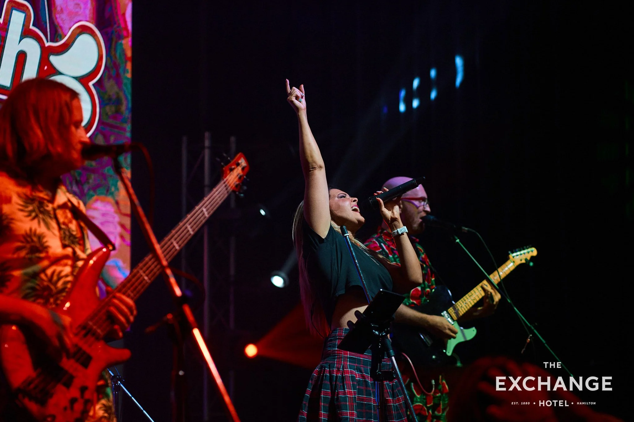 A group of musicians performing on stage at The Exchange Hotel in Hamilton. The woman in the center is singing into a microphone with her arm raised, wearing a black top and plaid skirt. Other musicians are playing guitars, dressed in colorful patterned shirts. The stage is dark with some blue lighting effects.