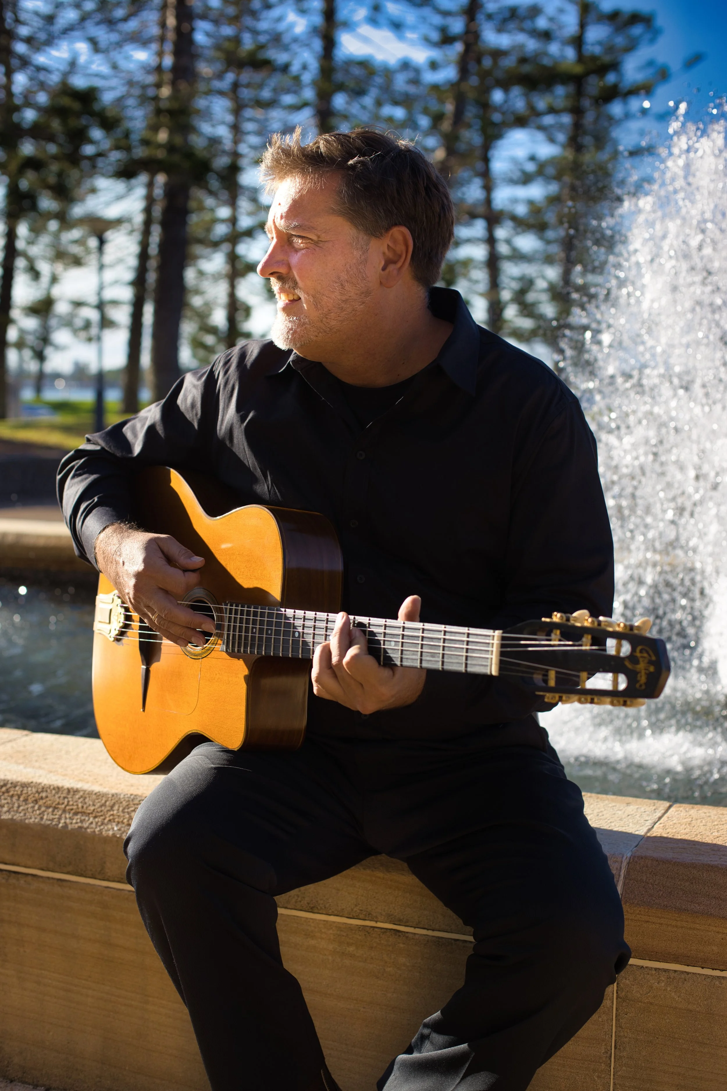 A man playing an acoustic guitar outdoors near a fountain and trees during the daytime.