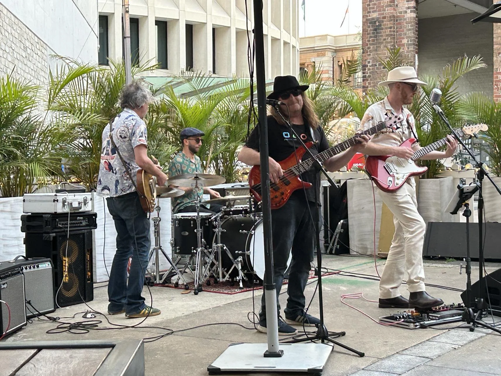 A four-member band performing outdoors, with palm plants in the background and brick buildings behind them. One musician plays bass guitar, another plays electric guitar, a third plays drums, and the fourth also plays guitar. They are dressed in casu
