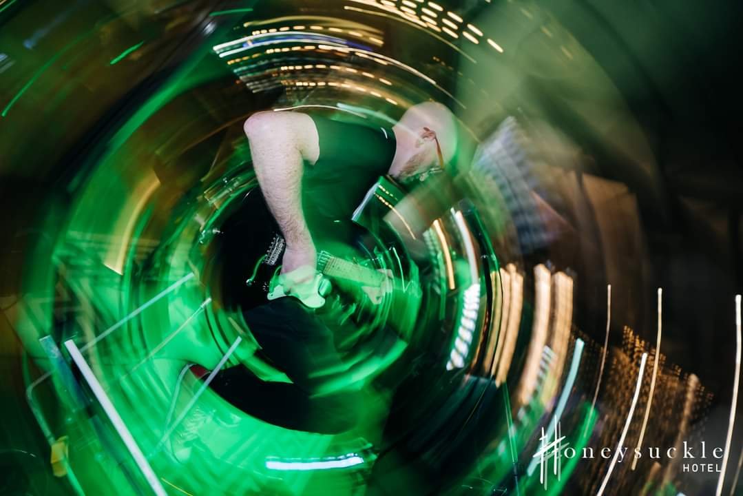 A musician playing an electric guitar on stage with green and yellow stage lighting, captured with a fisheye or swirl lens effect at Honeysuckle Hotel.