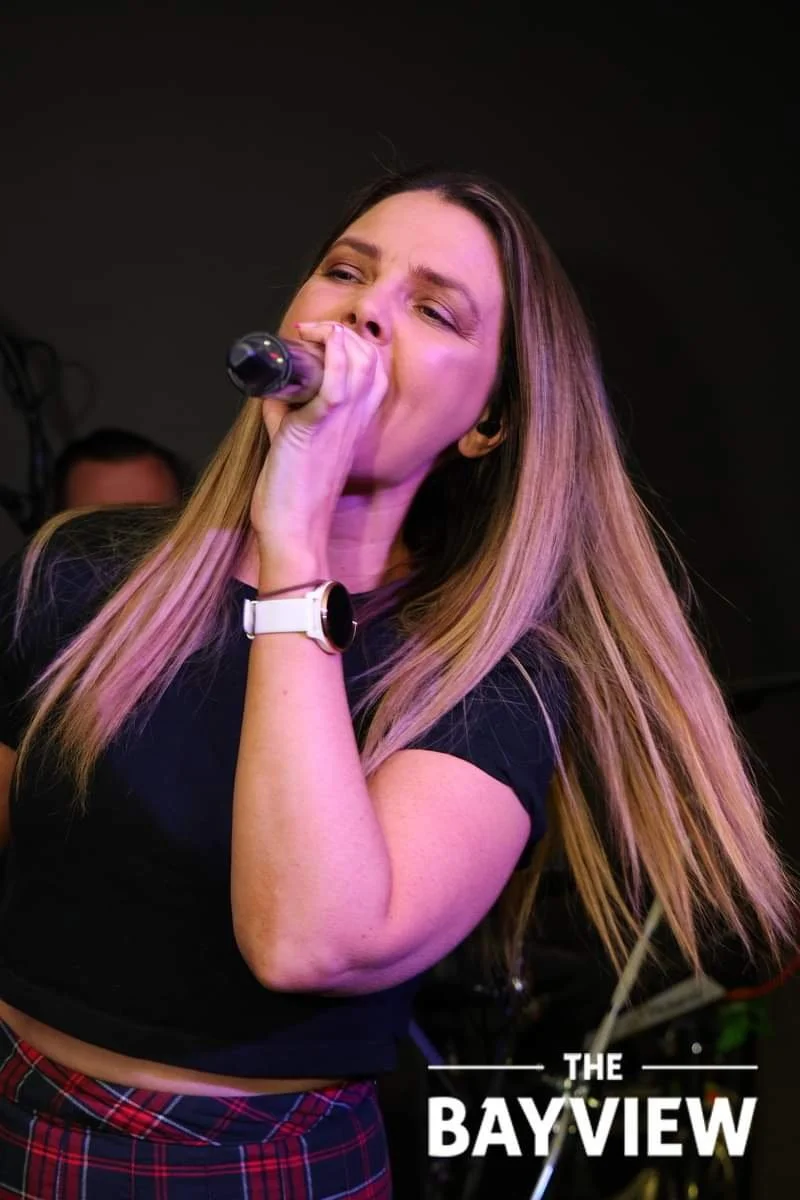 A woman with long blonde hair singing into a microphone at a venue called The Bayview.