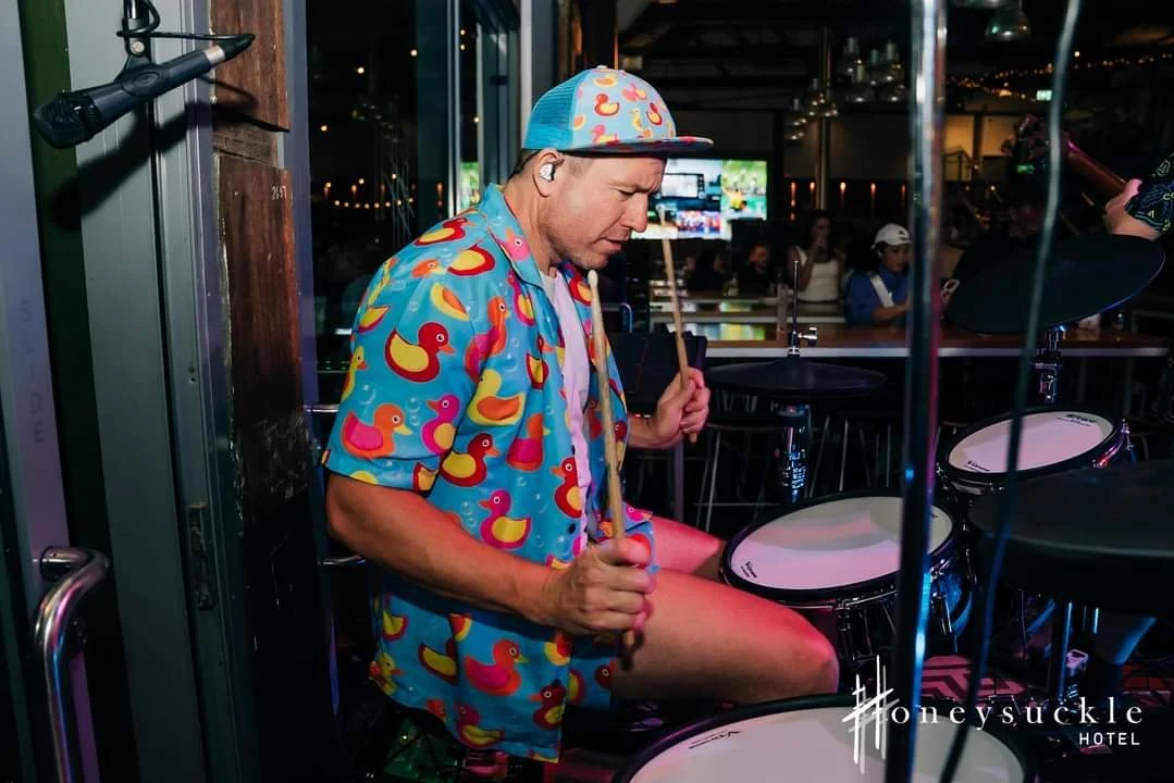 A man wearing a colorful shirt and a matching cap playing electronic drums in a bar or club at night, with people and a TV screen in the background.