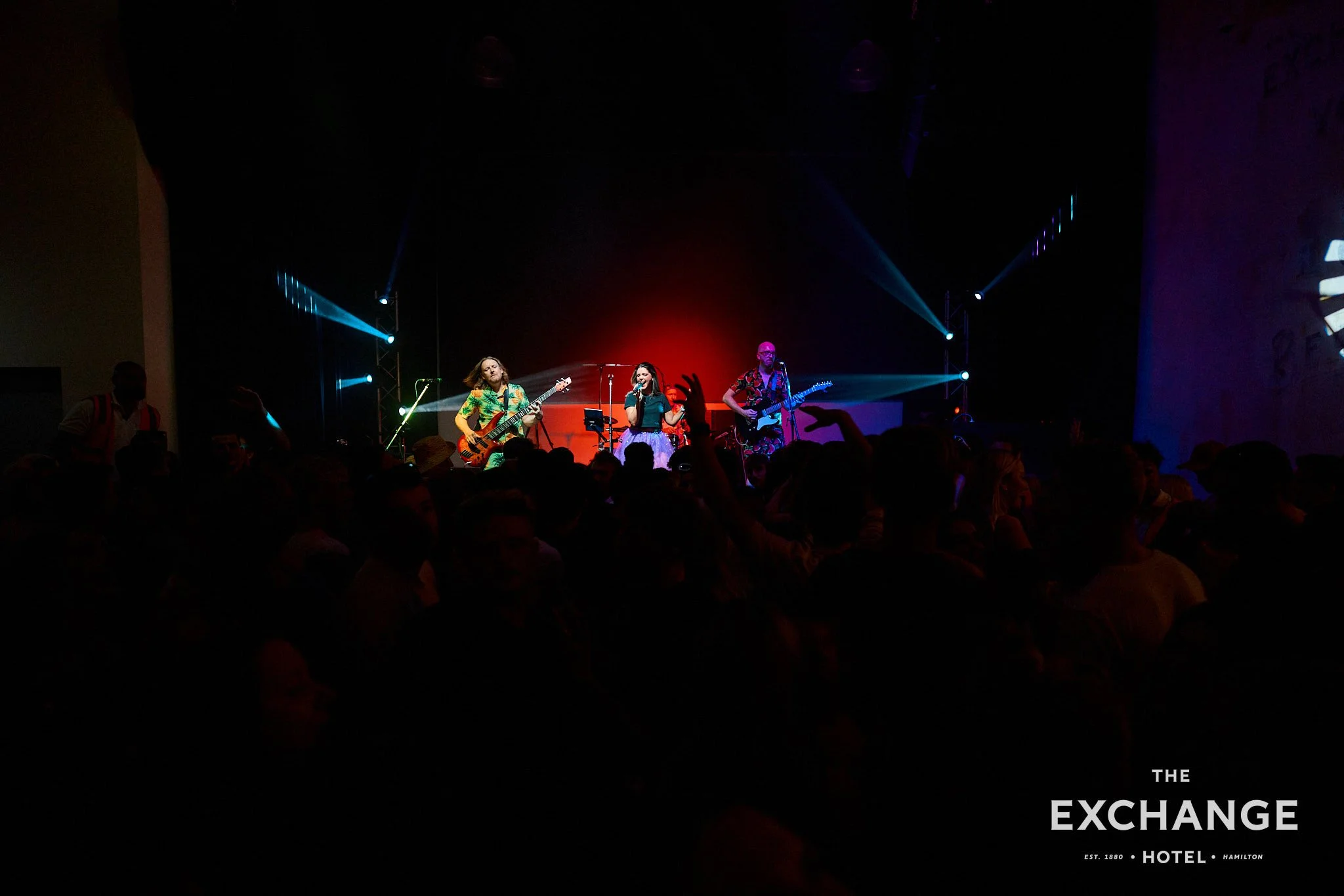 A live music performance at The Exchange Hotel in Hamilton with musicians playing guitars and singing, illuminated by stage lights in front of an audience.