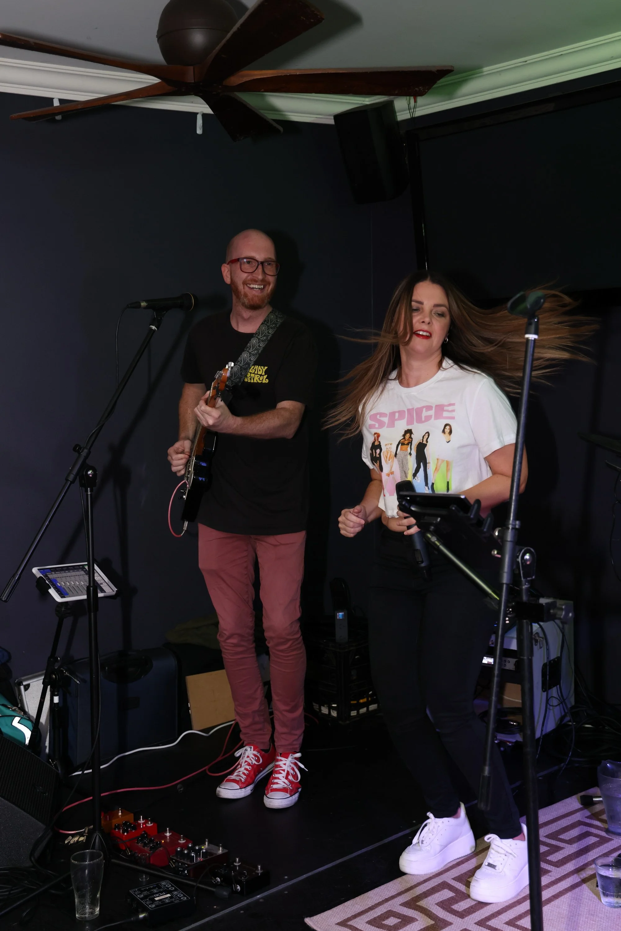 Two musicians perform indoors, with one playing an electric guitar and the other singing into a microphone, surrounded by audio equipment.
