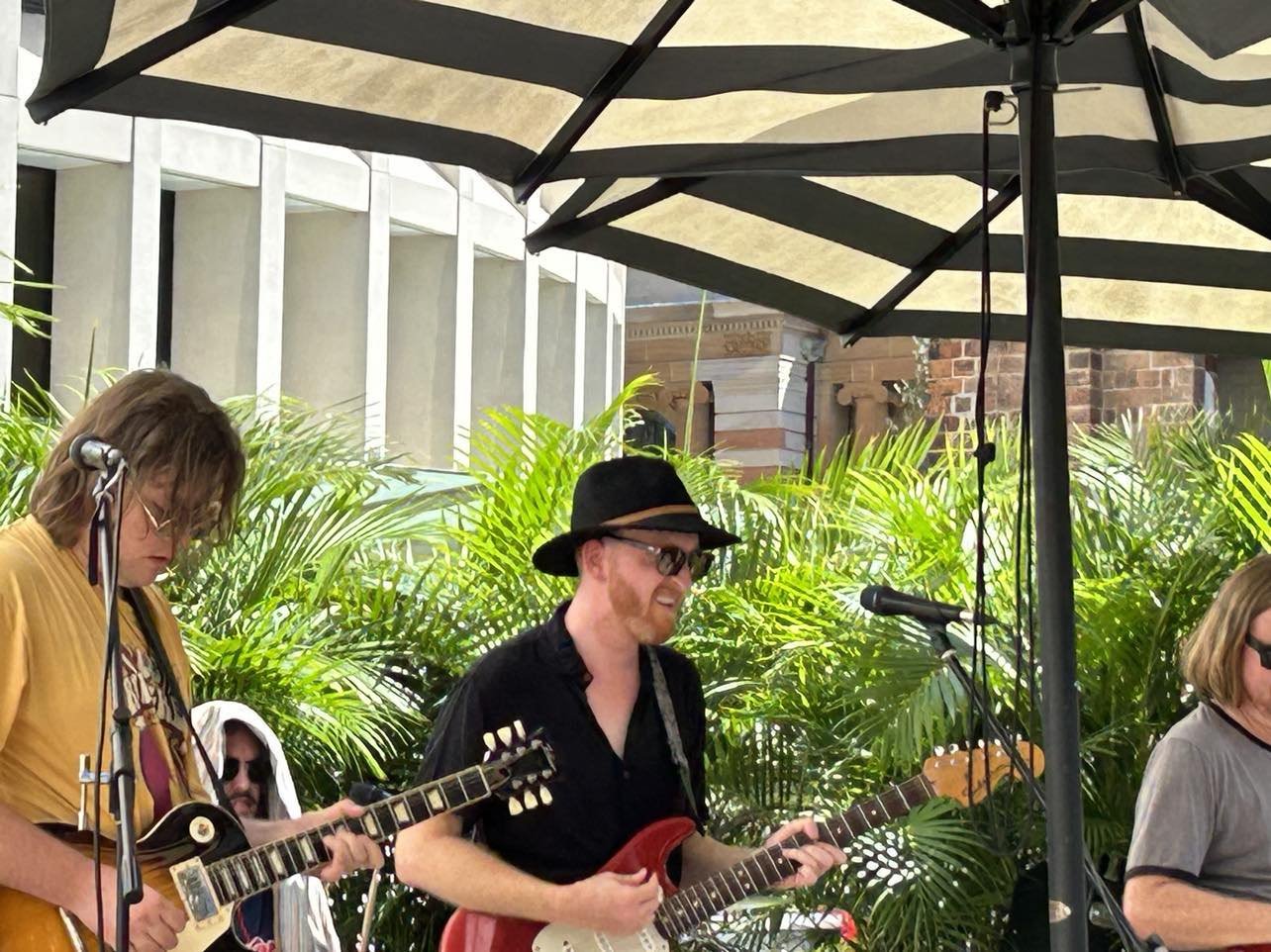 A group of musicians playing guitars outdoors under large patio umbrellas with green foliage in the background.