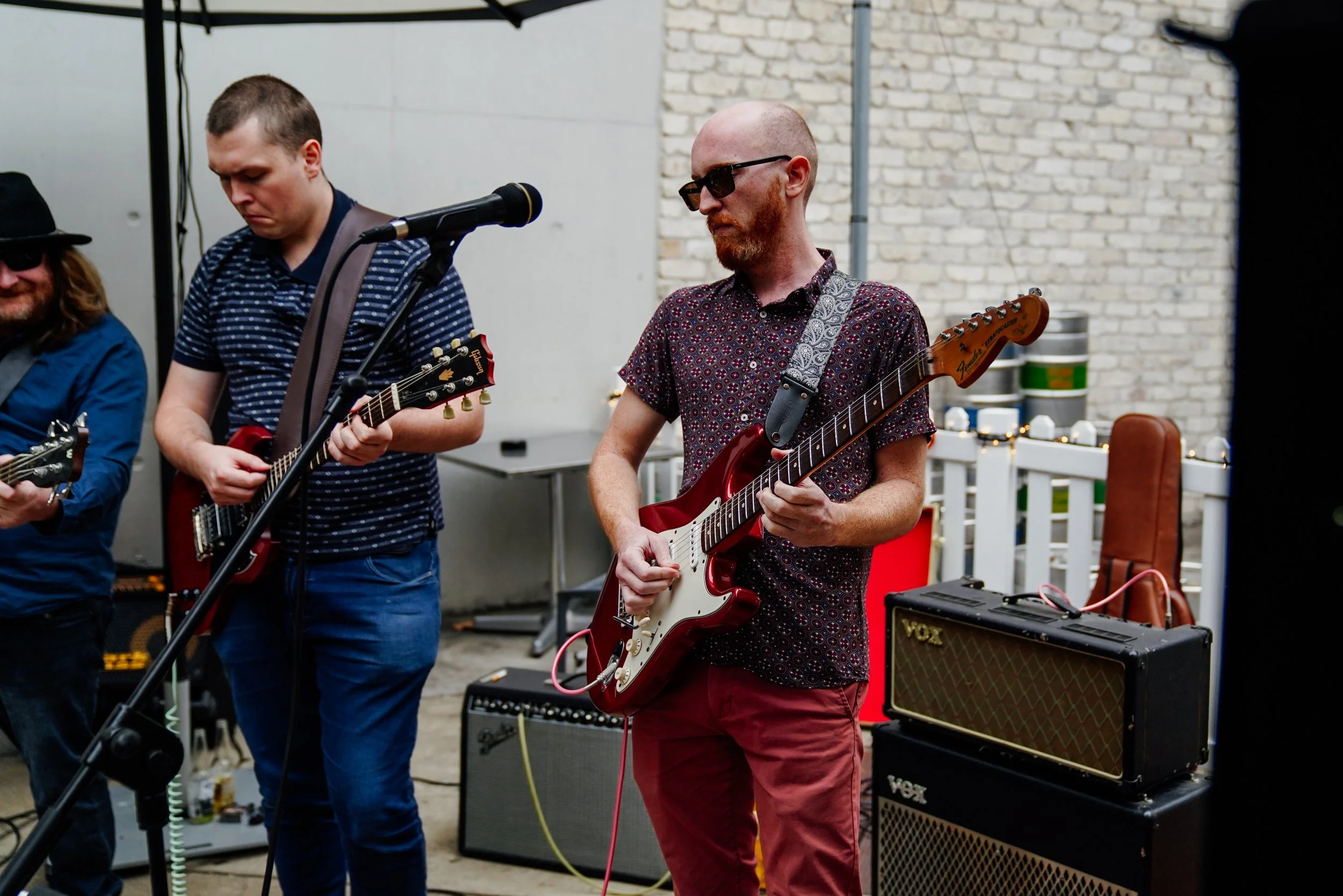 Three musicians performing with electric guitars, one man with a red guitar, another man with a black guitar, and a man on the left with a black hat and glasses. They are outdoors against a background of brick and concrete walls, with musical equipme