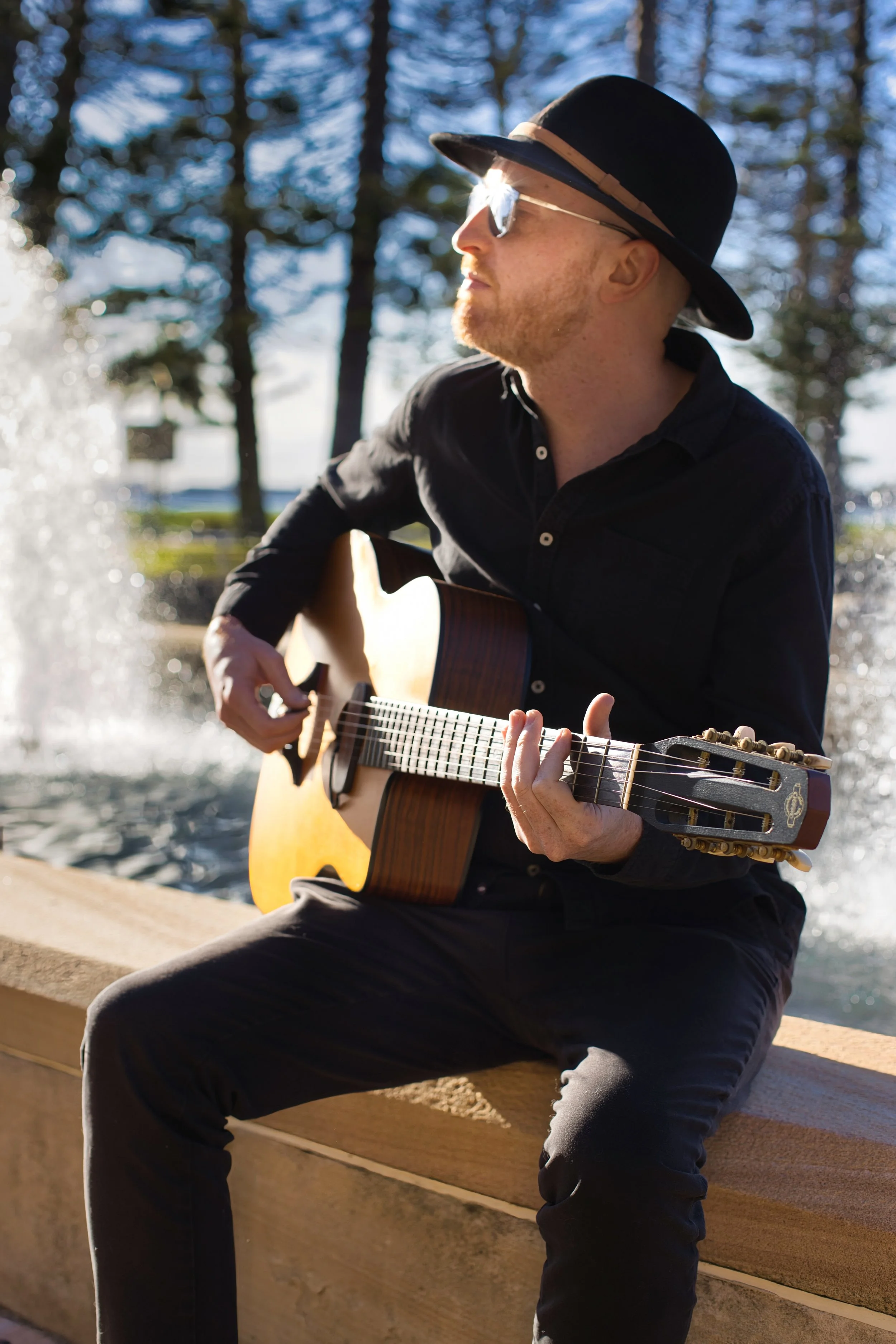 A man sitting on a stone bench outdoors, playing an acoustic guitar, wearing sunglasses and a black wide-brimmed hat, with trees and a water fountain in the background.