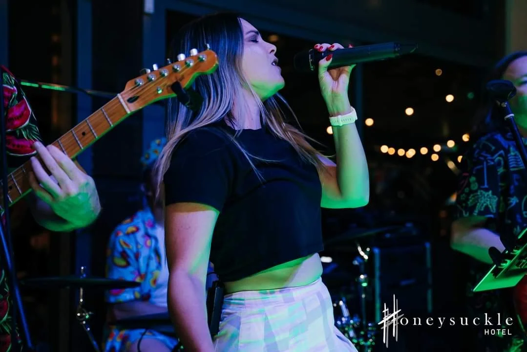 Female singer performing with a microphone at a music venue with musicians playing instruments in the background
