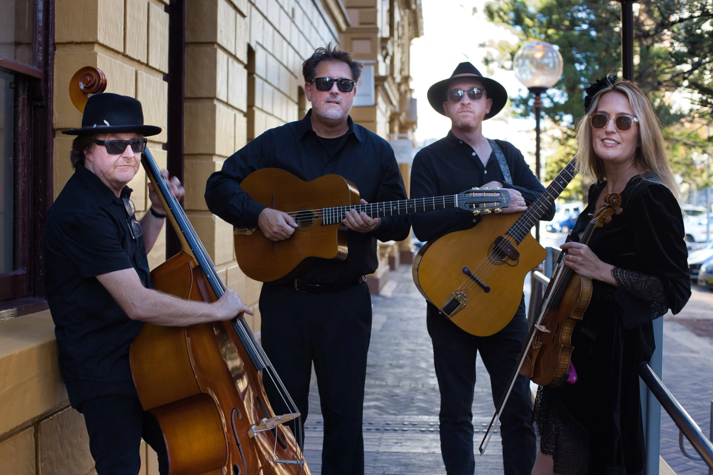 A group of four street musicians, three men and one woman, playing guitars, a violin, and a double bass on a city sidewalk during daytime.
