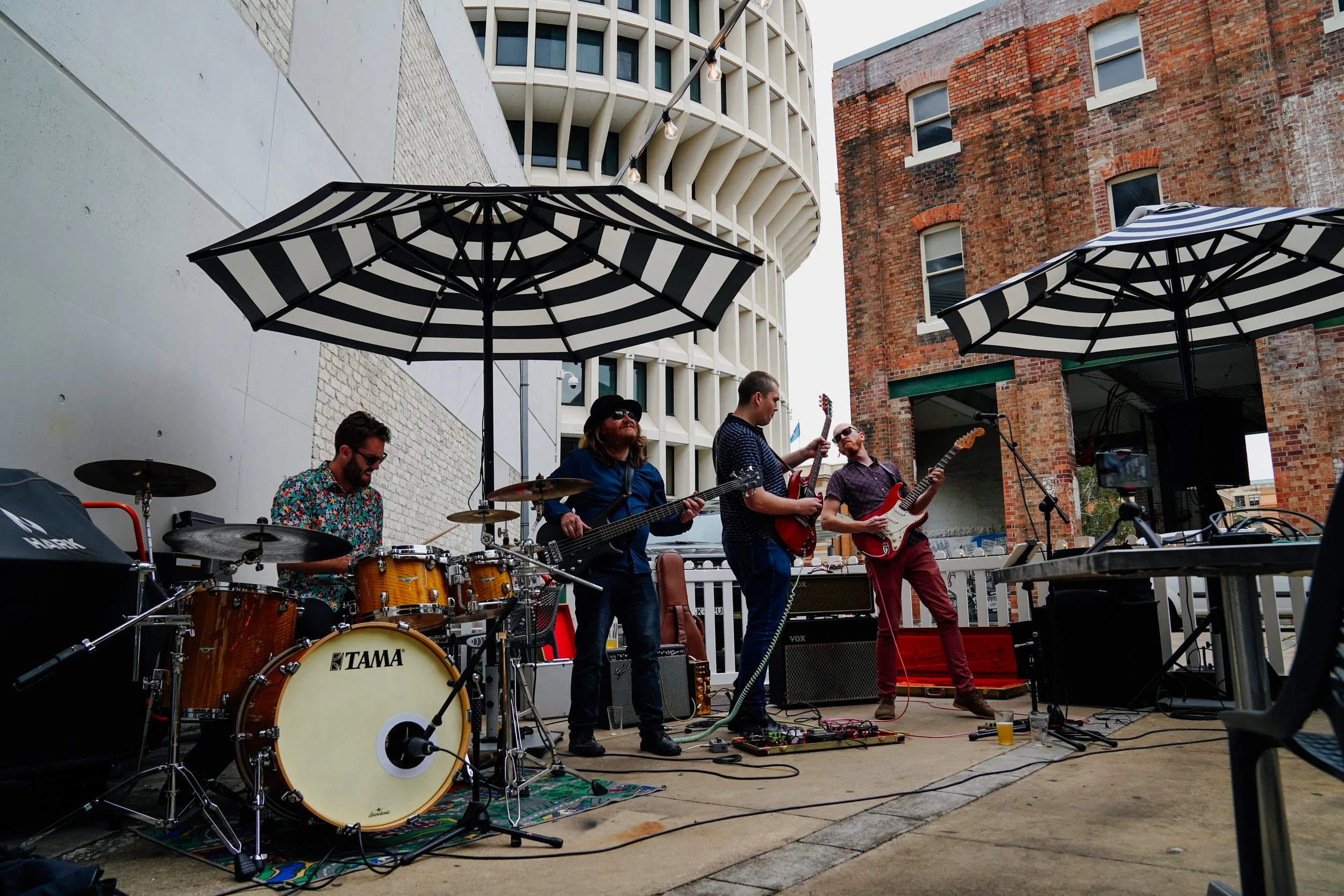 A band performing outdoors on a small stage with umbrellas overhead, on a city street with brick and modern buildings behind them.