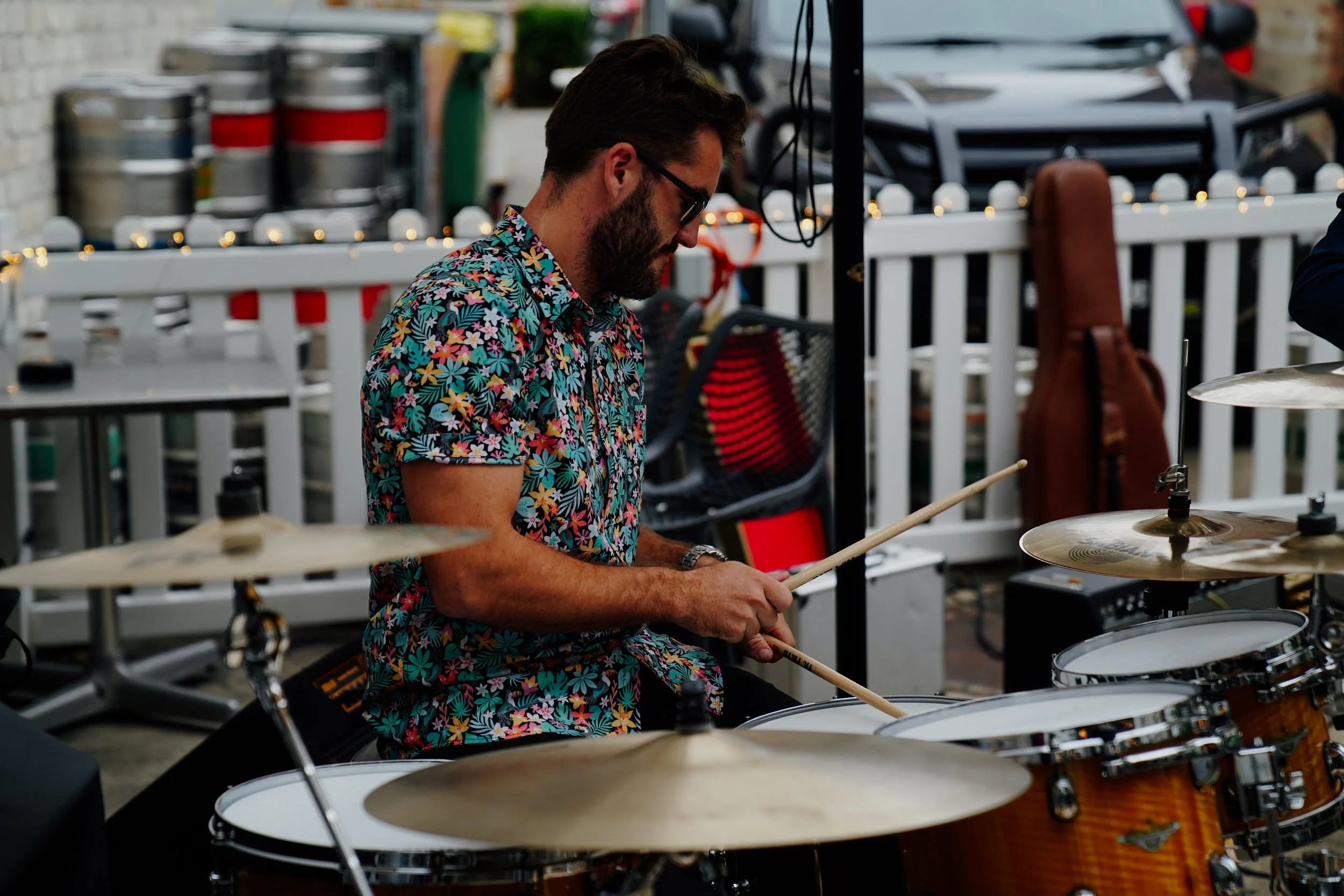A man with dark hair, beard, sunglasses, and a colorful floral shirt playing drums at an outdoor event.