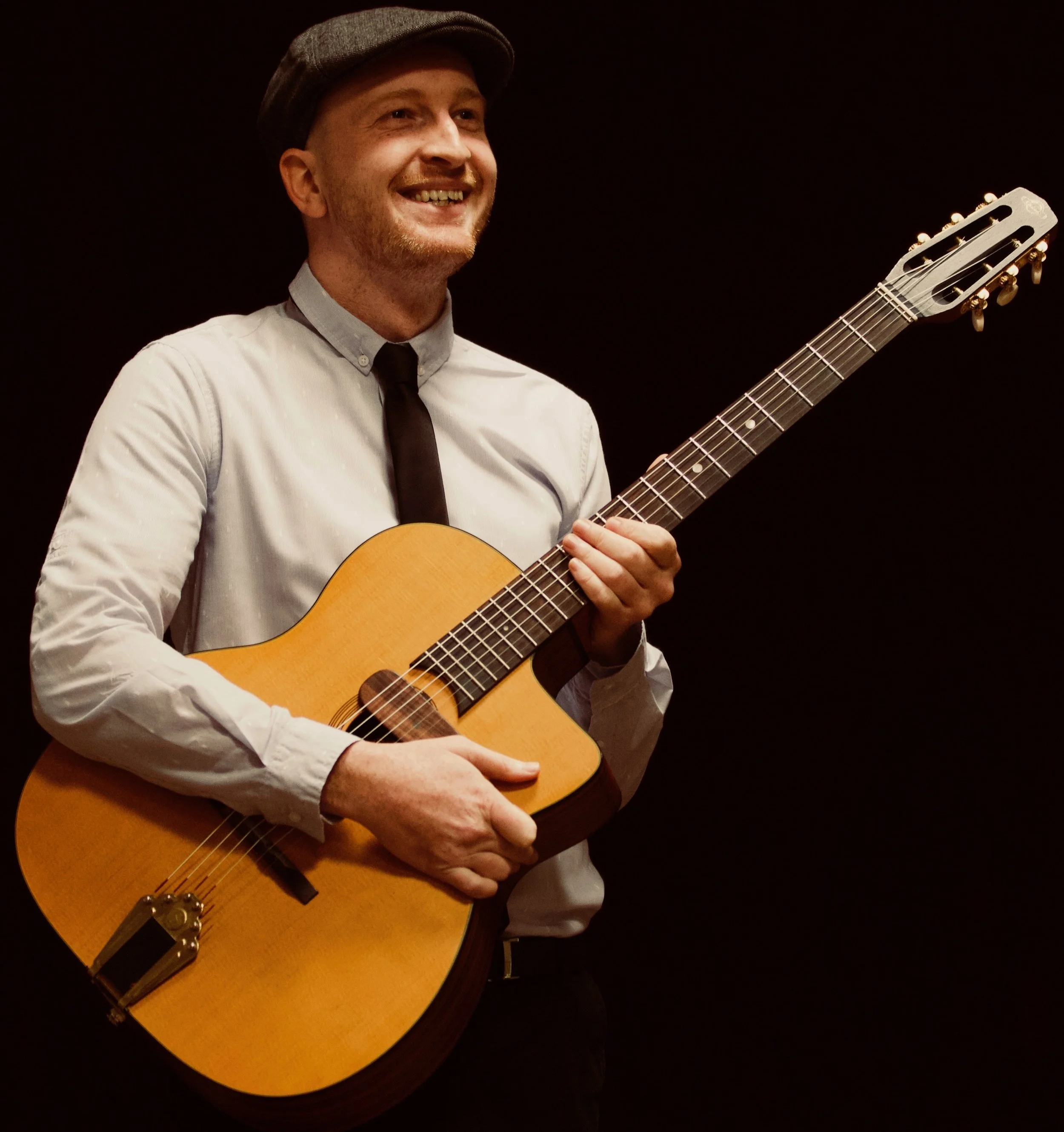 A man smiling and holding an acoustic guitar against a dark background. He is wearing a light-colored shirt, a dark tie, and a hat.