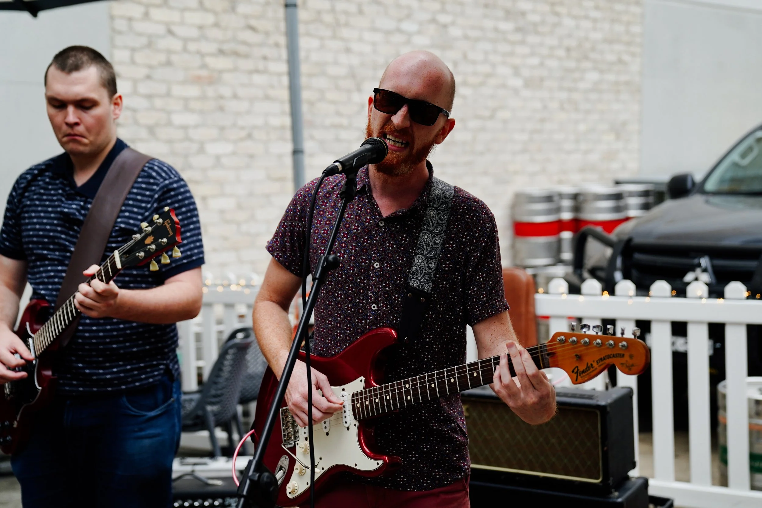 Two men playing electric guitars outdoors in front of a brick wall and white picket fence. The man on the right has a shaved head, sunglasses, a patterned shirt, and is singing into a microphone. The man on the left has short dark hair, a striped shi