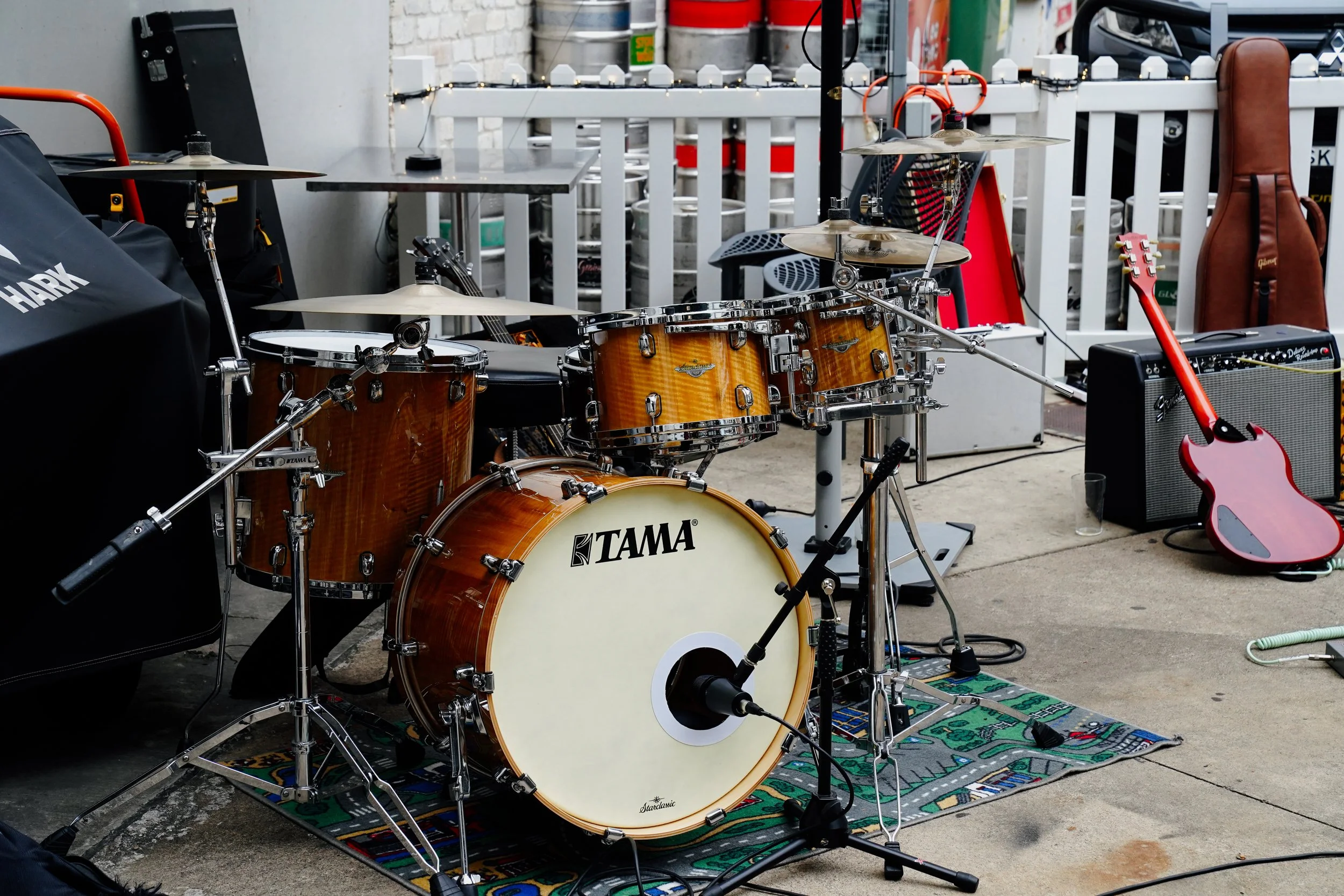 A drum set on a colorful patterned rug outside, with a bass drum labeled TAMA, surrounded by various musical equipment including an electric guitar, amplifier, and microphone, set against a white picket fence and barrels.