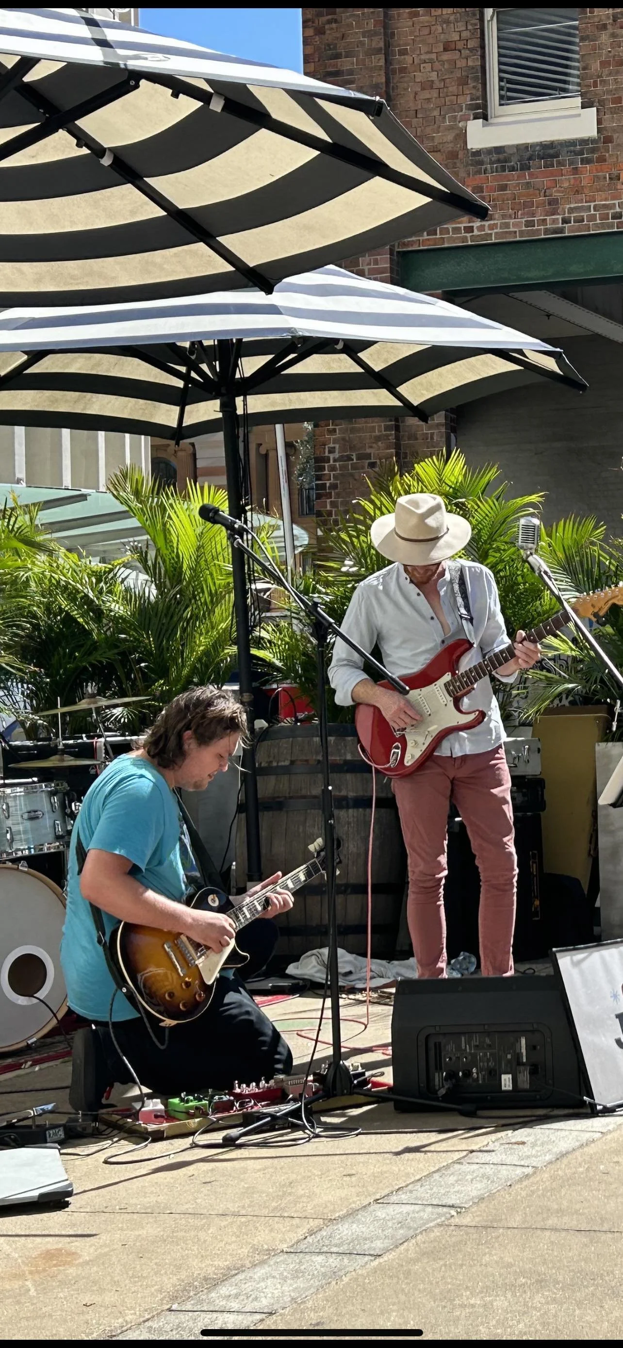 Two musicians playing electric guitars outdoors under large umbrellas, with lush green plants, brick buildings, and a window in the background.