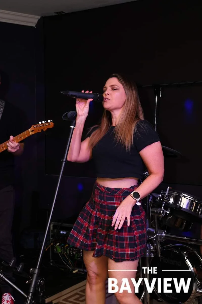 A woman singing into a microphone on stage, wearing a black crop top and a red plaid skirt, with musical instruments and equipment around her.