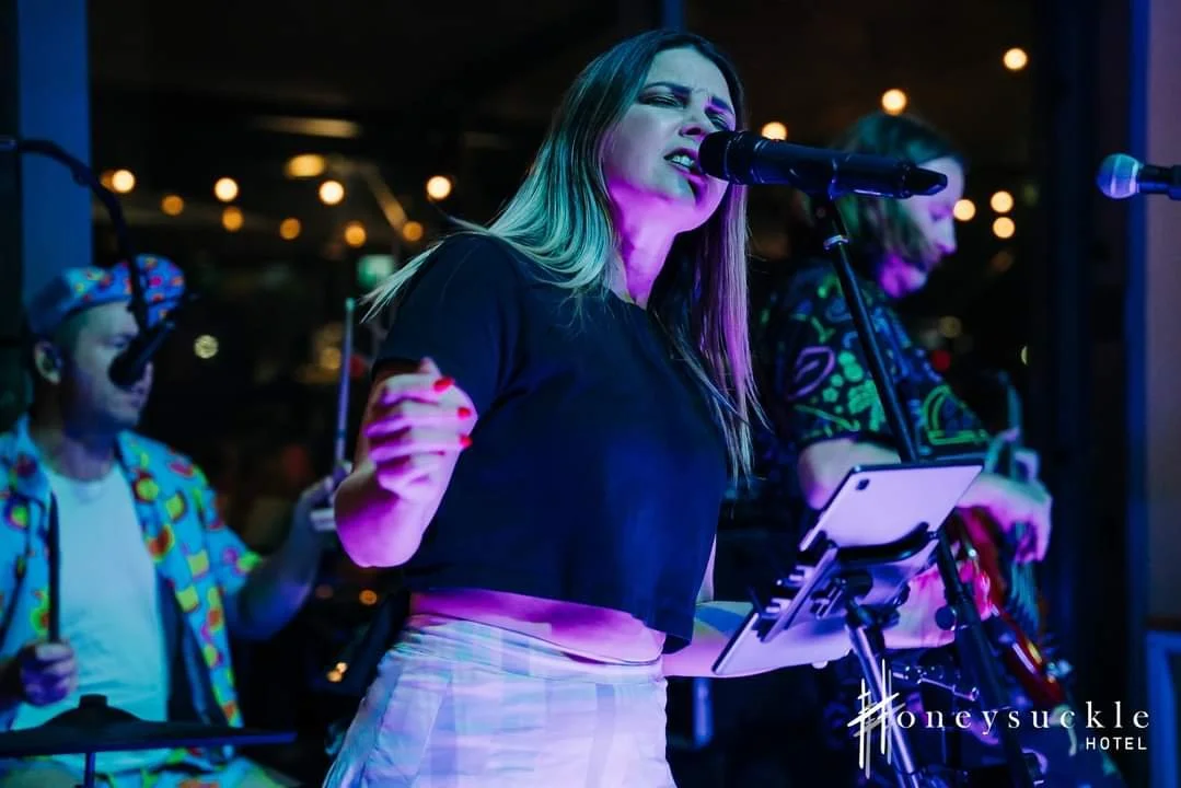 A woman singing into a microphone on stage with two musicians in the background at a hotel event.