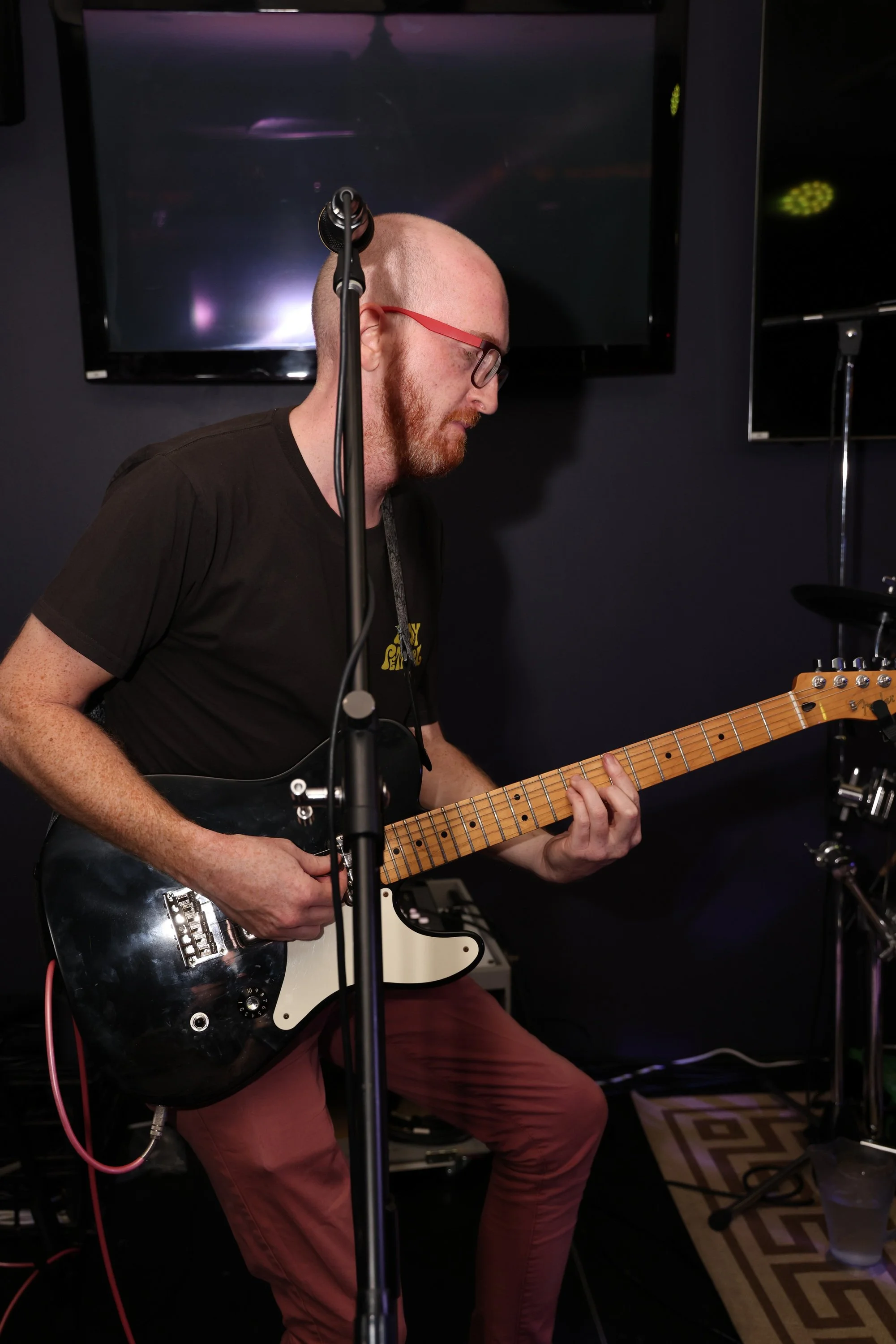 A man with a shaved head and red beard wearing glasses and a black t-shirt playing an electric guitar during a live performance, with a dark background and television screens.