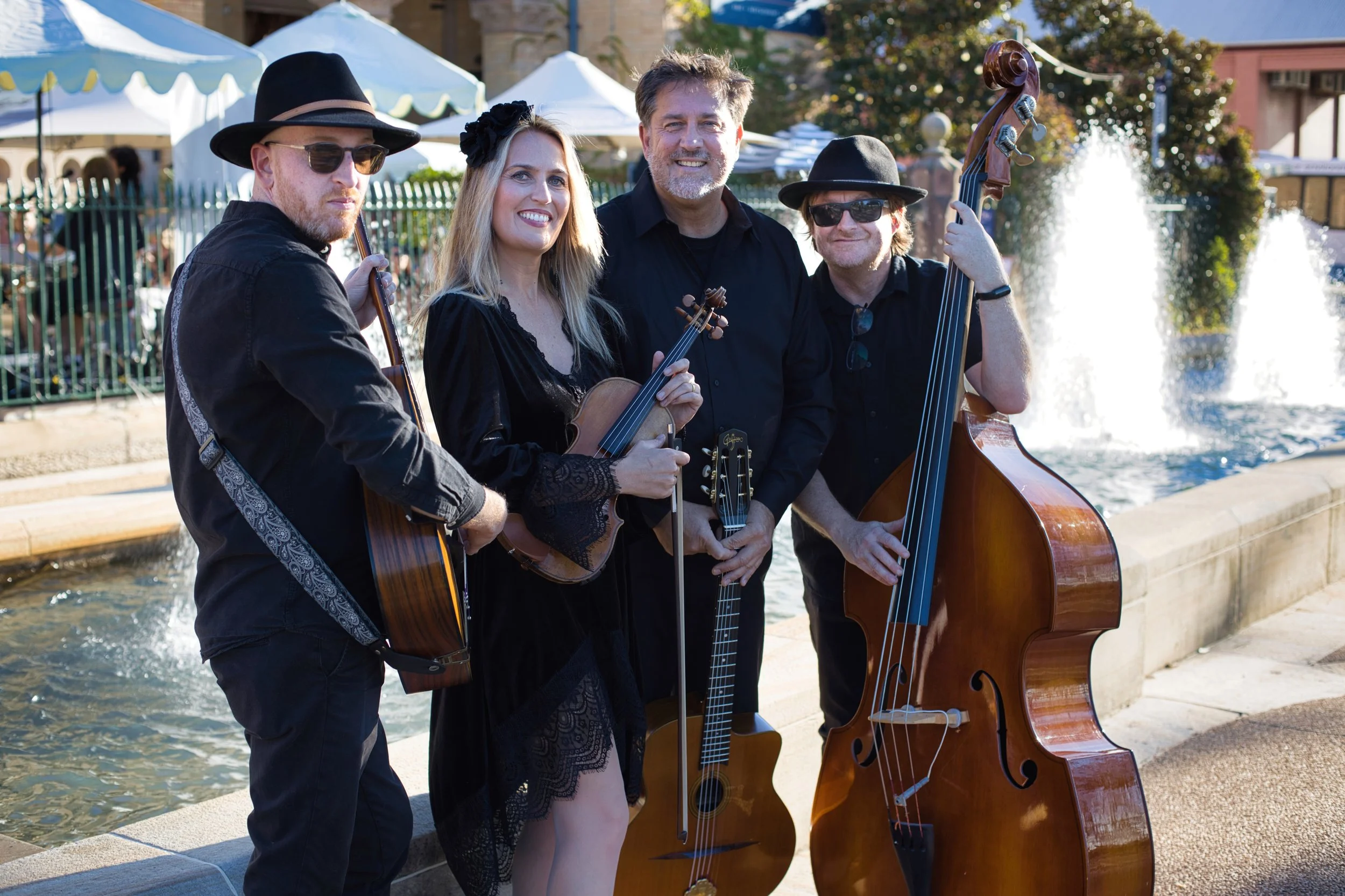 A group of four musicians outdoors near a fountain, holding musical instruments including an acoustic guitar, violin, and upright bass, dressed in black, smiling at the camera, with umbrellas and trees in the background.