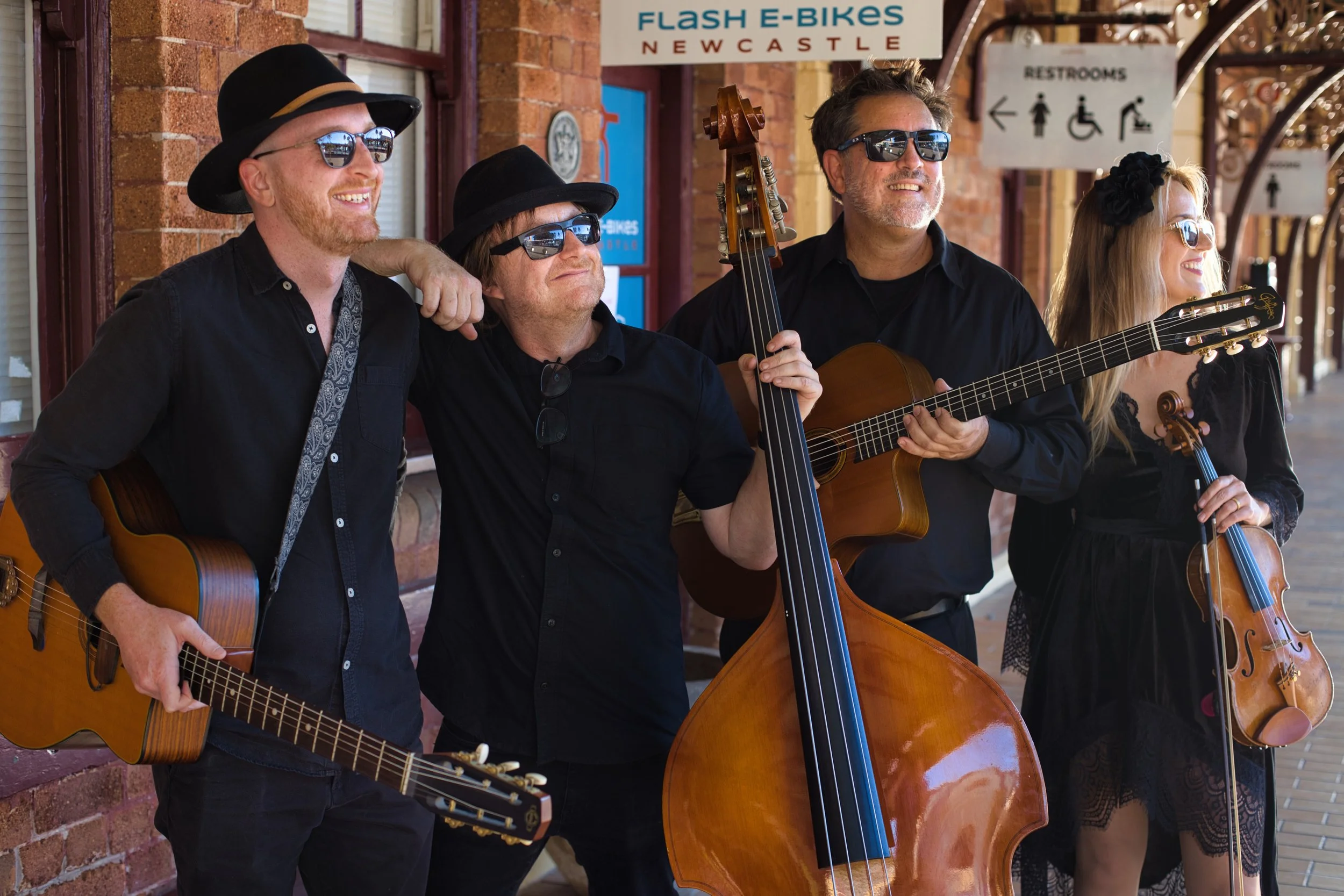 Four musicians with guitars standing outside a brick building, wearing sunglasses and black clothing, smiling and enjoying music, with signs overhead for restrooms and a nearby sign that reads "Flash E-Bikes Newcastle."
