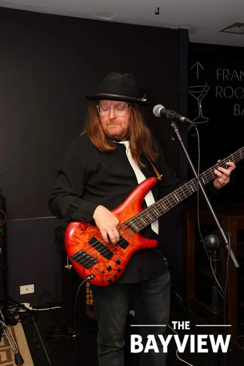 A man wearing a black hat, black shirt, and white tie playing a red electric bass guitar in a dimly lit room with a microphone nearby.