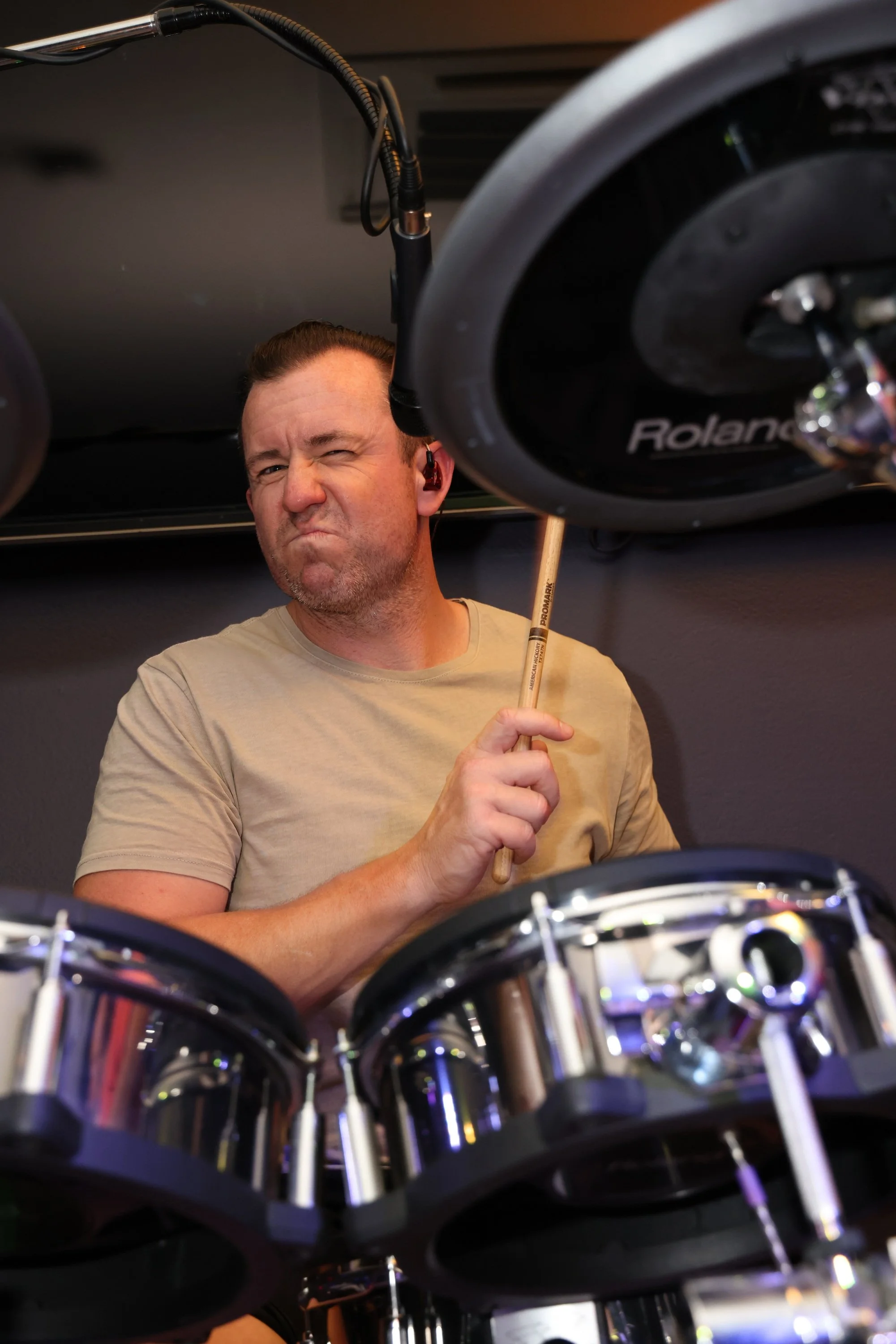 A man making a silly face, winking with one eye and grimacing, surrounded by musical instruments including a drum set, with a microphone overhead.
