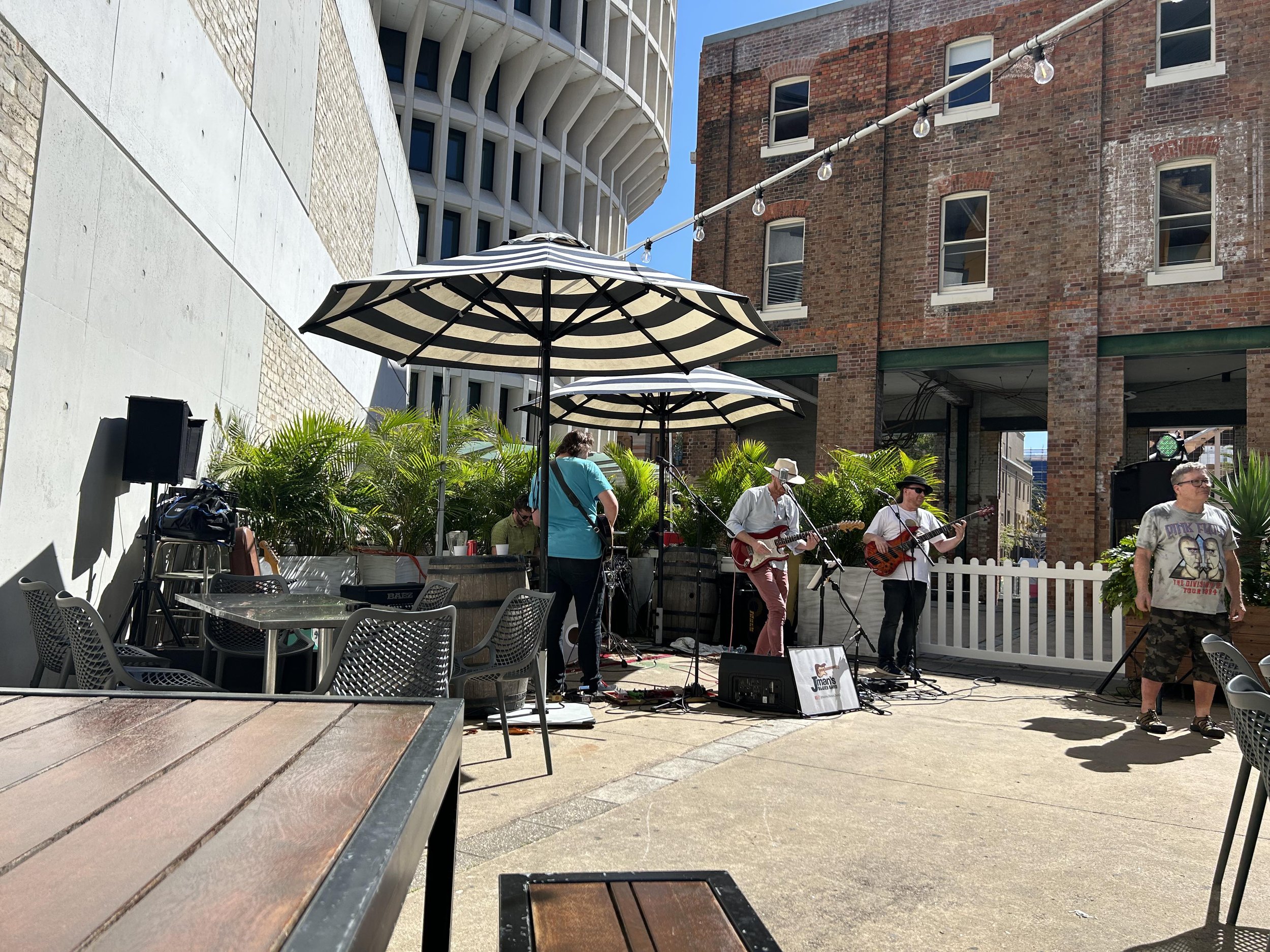 A live outdoor music performance with a band playing on a stage, featuring guitars and a keyboard, under black and white striped umbrellas on a city street with brick and concrete buildings, greenery, and side tables and chairs in the foreground.